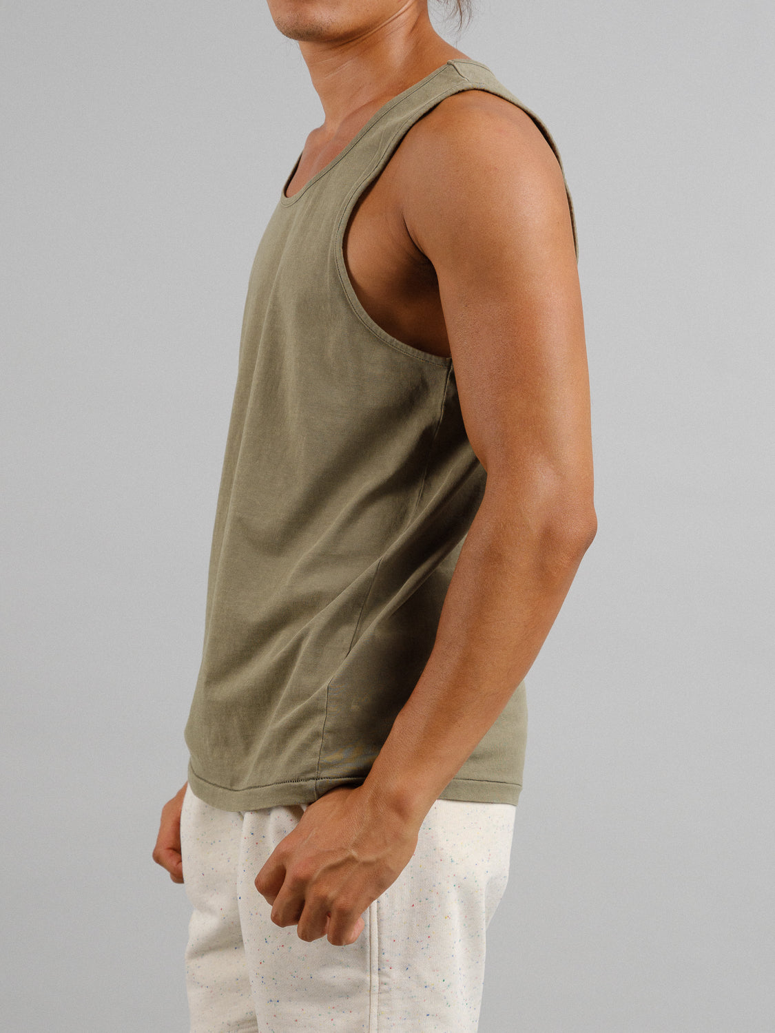 A person models the ID Menswear Settler Snow Vintage Wash Tank Top in sleeveless olive green, paired with light shorts against a plain gray background. The lightweight tank top underscores their upper body silhouette from the side, omitting their face.