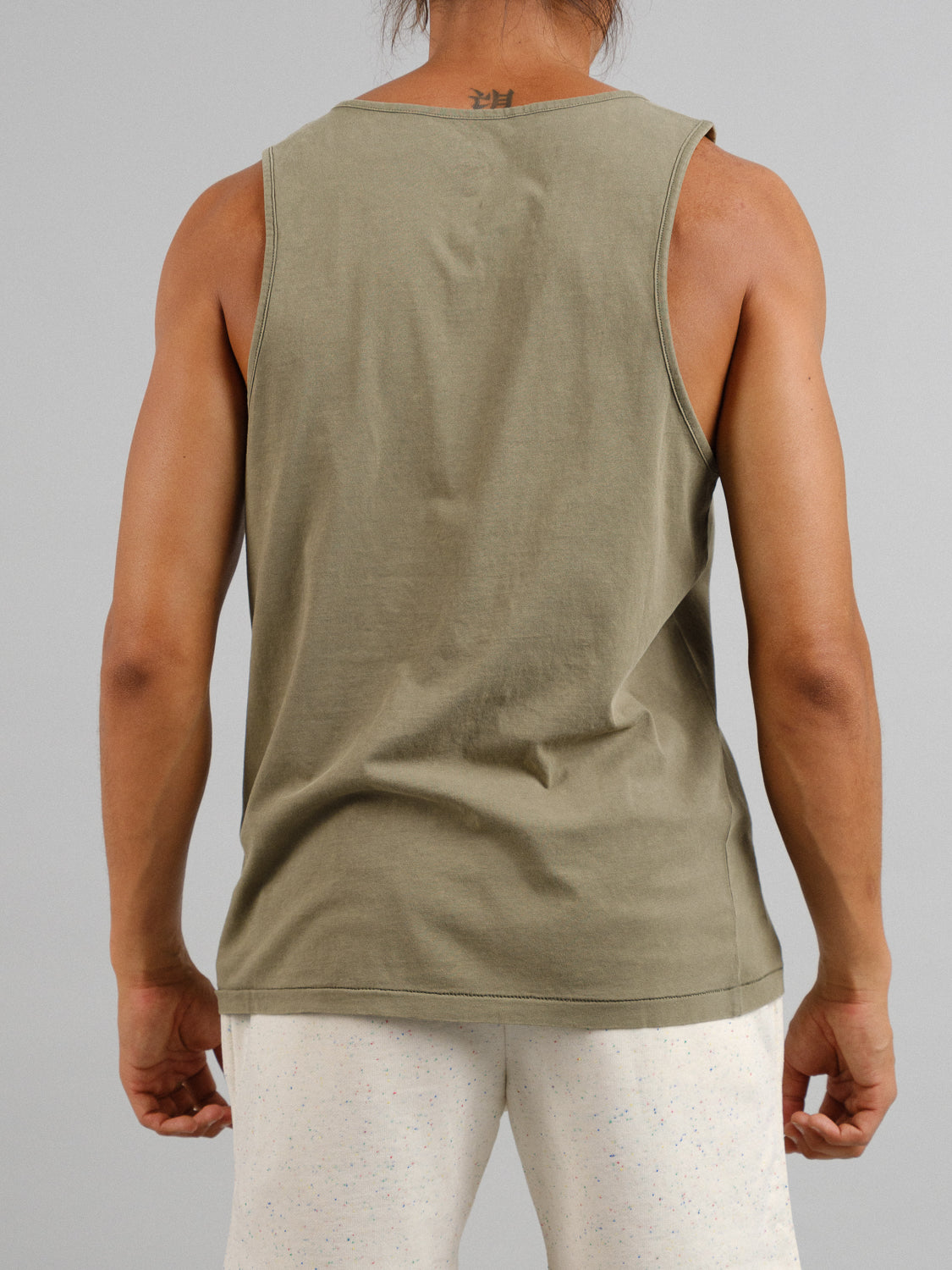 Seen from behind, a person with long hair tied back wears an olive green Settler Snow Vintage Wash Tank Top by ID Menswear, styled with light speckled shorts. The plain gray background accentuates the ensembles vintage appeal.