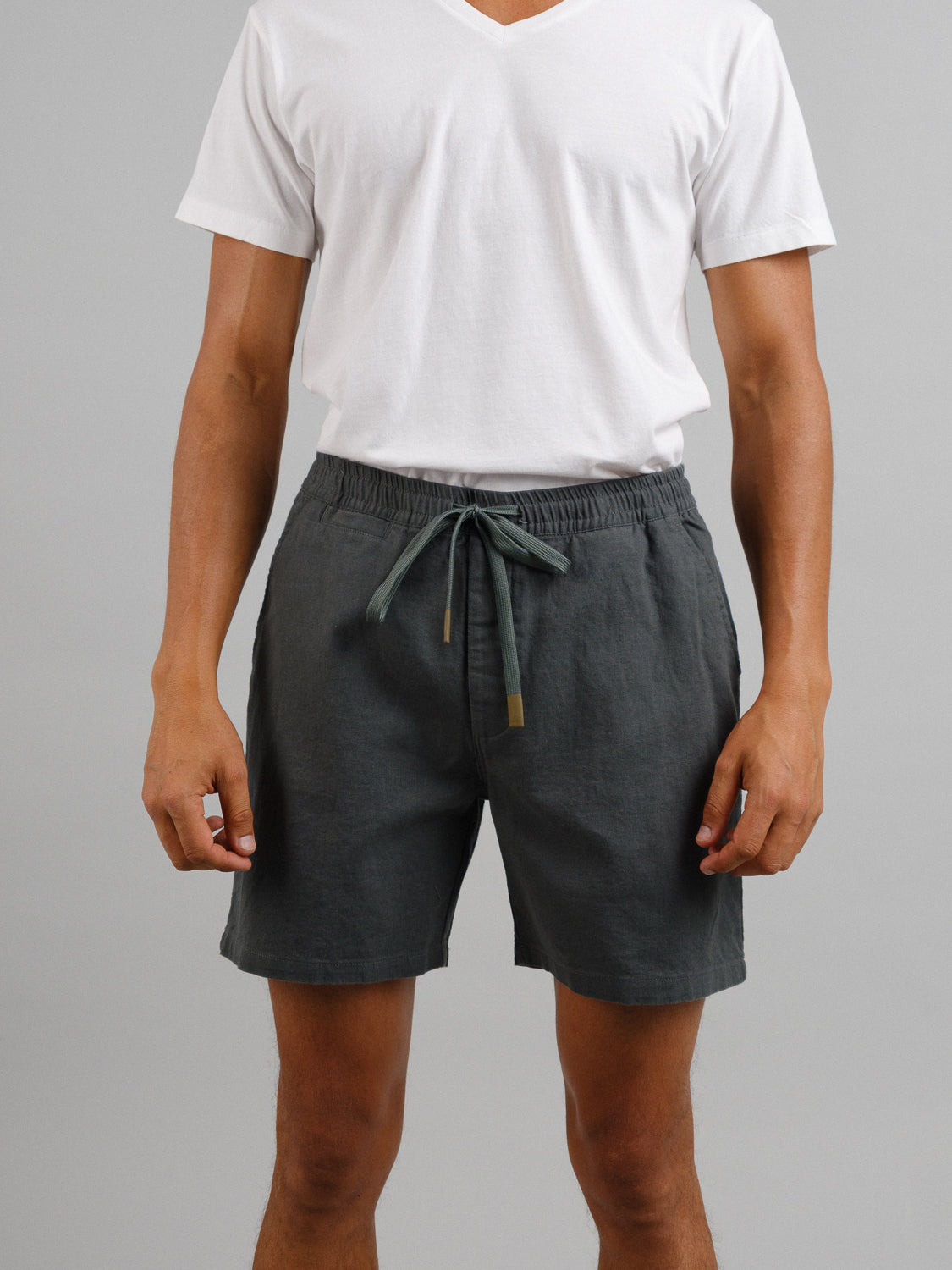 A person wearing a plain white V-neck T-shirt paired with ID Menswears Kotao Linen Blend Drawstring Short in dark gray stands against a minimalistic gray backdrop, highlighting the casual summer appeal of the outfit.
