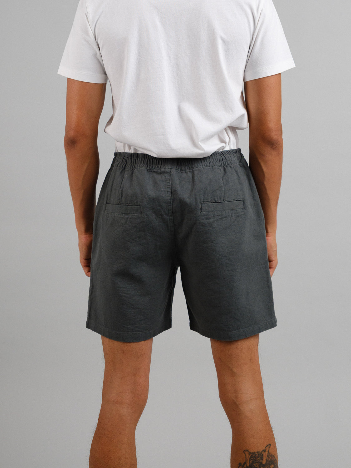 A rear view shows someone in a white T-shirt and ID Menswears Kotao Linen Blend Drawstring Shorts, featuring an elastic waistband. They display a tattoo on their left calf against a gray backdrop, ideal for warm weather outings.