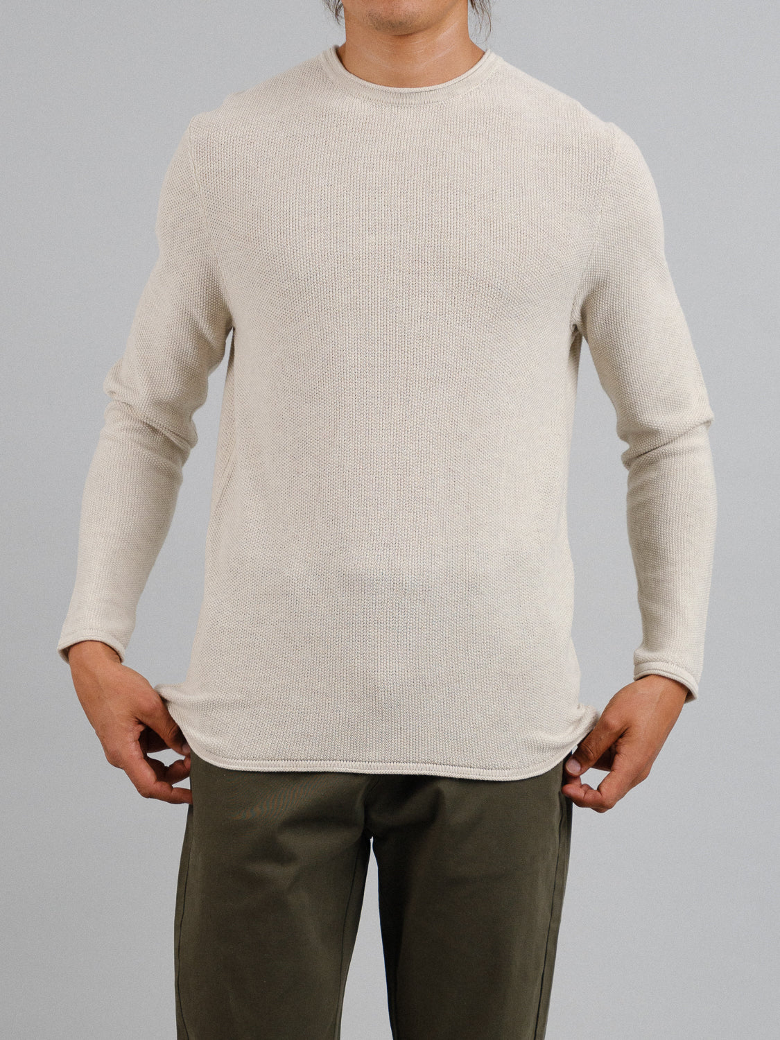 A person models the ID Menswear Madrid Rolled Neck Sweater Tee in light beige alongside olive green pants, set against a plain gray backdrop. The arms are slightly away from the body, with hands near the sides of the sweater, but the head is not visible.
