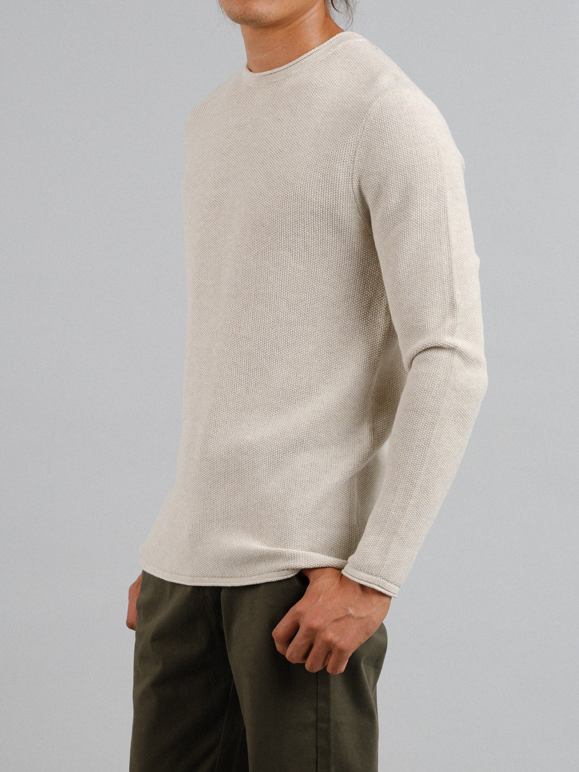 Against a gray background, a person stands wearing ID Menswears Madrid Rolled Neck Sweater Tee in light beige with a subtle mini popcorn knit texture. Dark green pants complete the outfit as their left hand rests on their hip.