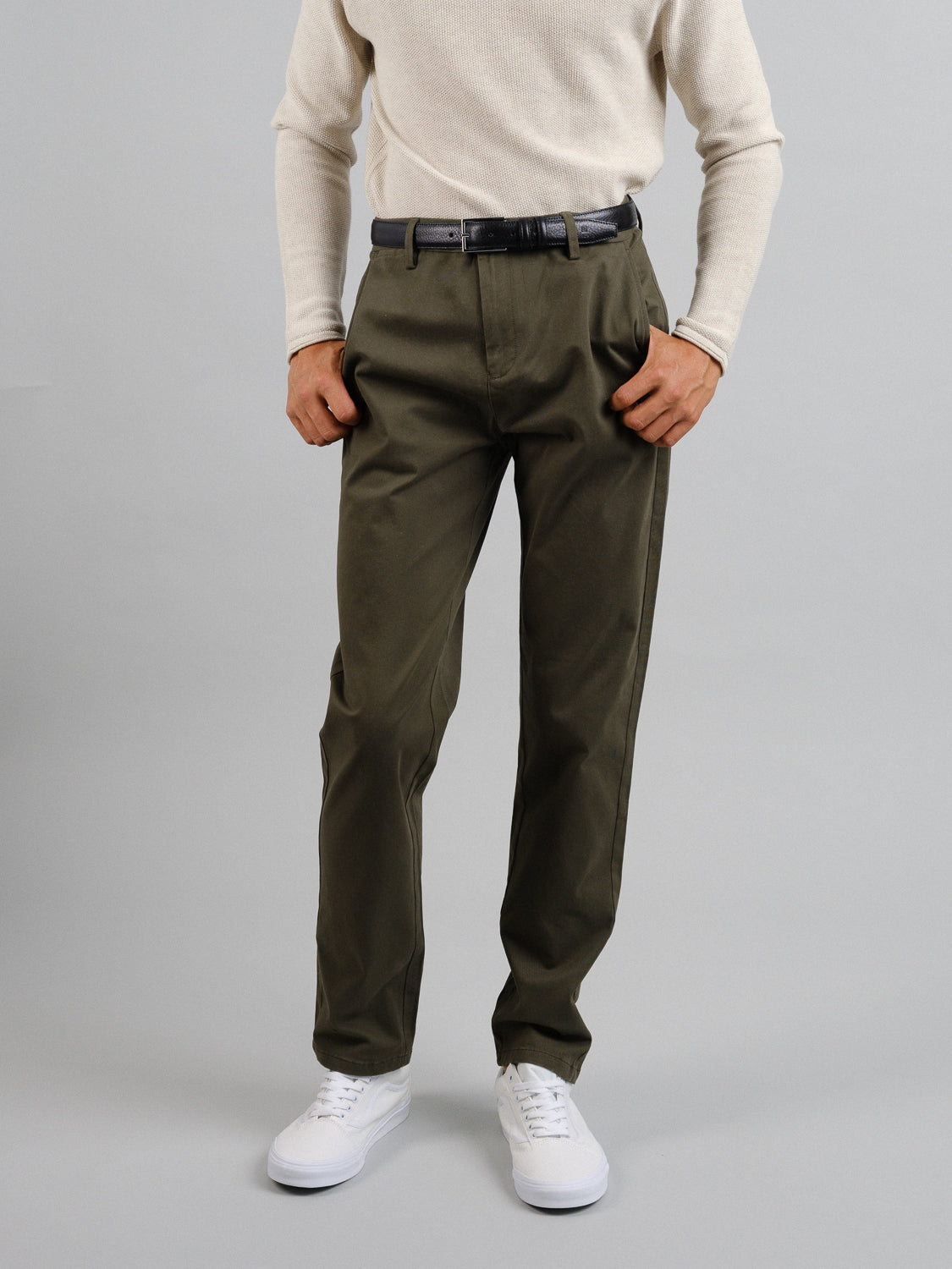 A person in olive green Bushwick Twill Stretch Chino trousers from ID Menswear, a light beige long-sleeve shirt, and white sneakers stands against a plain gray background with their hands in their front pockets, showcasing a black belt.