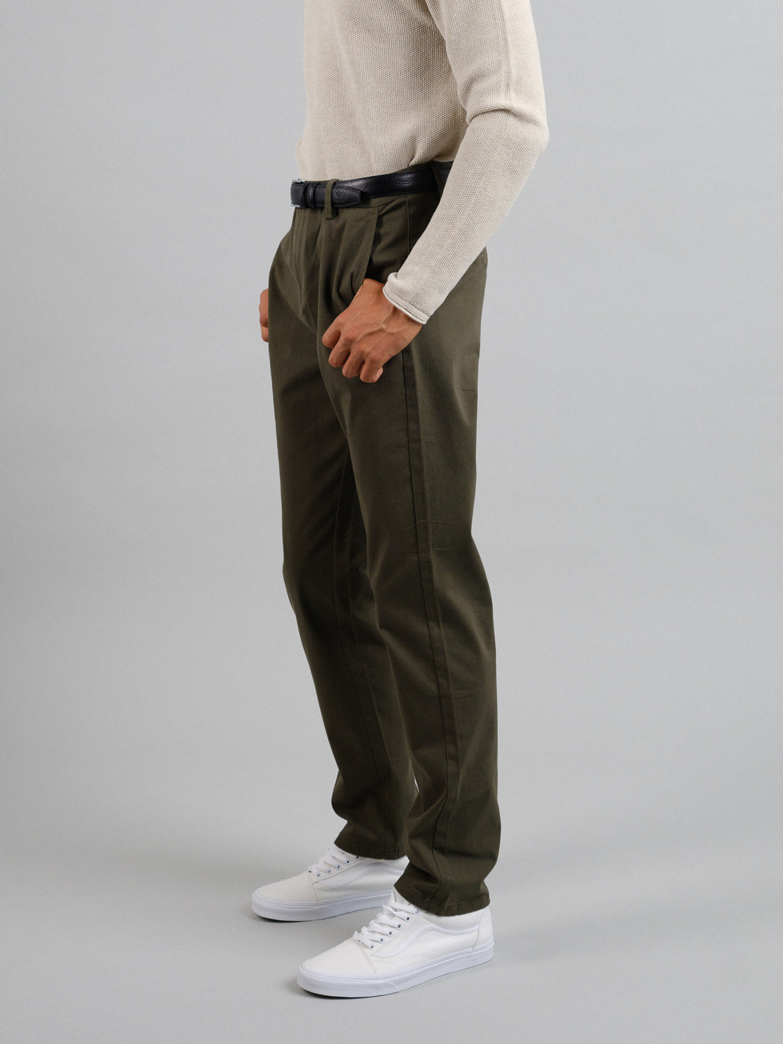 A person is wearing an ID Menswear cream long-sleeve shirt, Bushwick Twill Stretch Chino in dark olive green, and white sneakers. Theyre standing sideways, cropped from the shoulders down against a plain gray background.