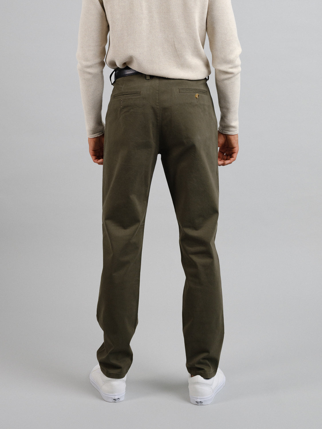 A person in an ID Menswear beige sweater and Bushwick Twill Stretch Chino trousers stands with their back to the camera, wearing white sneakers against a plain gray background.
