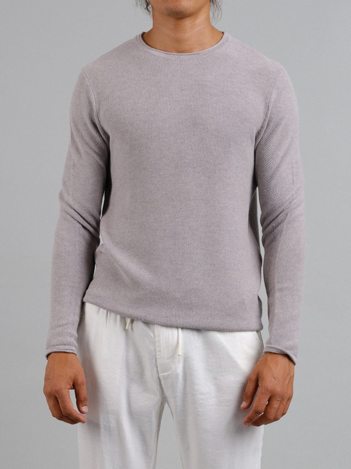 A person models the ID Menswear Madrid Rolled Neck Sweater Tee, a light gray, form-fitting sweater with a mini popcorn knit and soft rolled neck. They wear white drawstring pants and stand against a plain gray background, their hands relaxed at their sides.