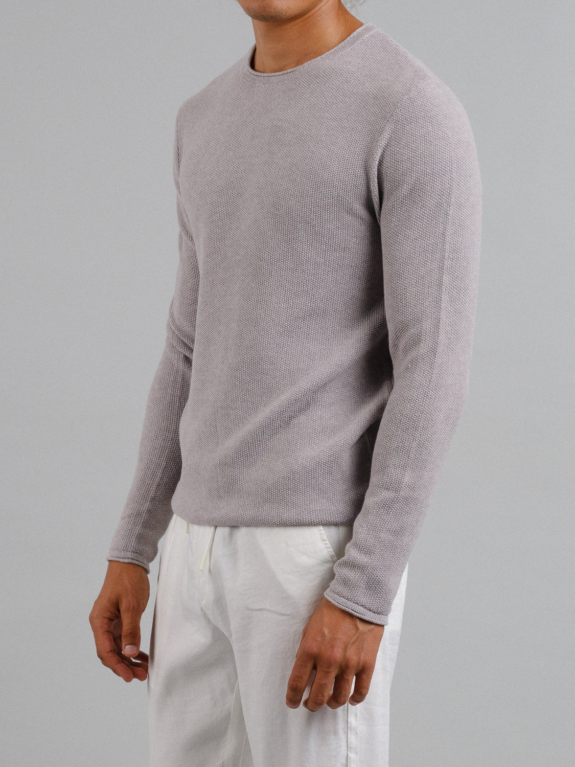 An individual models the Madrid Rolled Neck Sweater Tee by ID Menswear, showcasing its light gray mini popcorn knit and rolling neck. Paired with white pants, they stand against a plain gray background, angled to show both the side and front views. The head is out of frame.