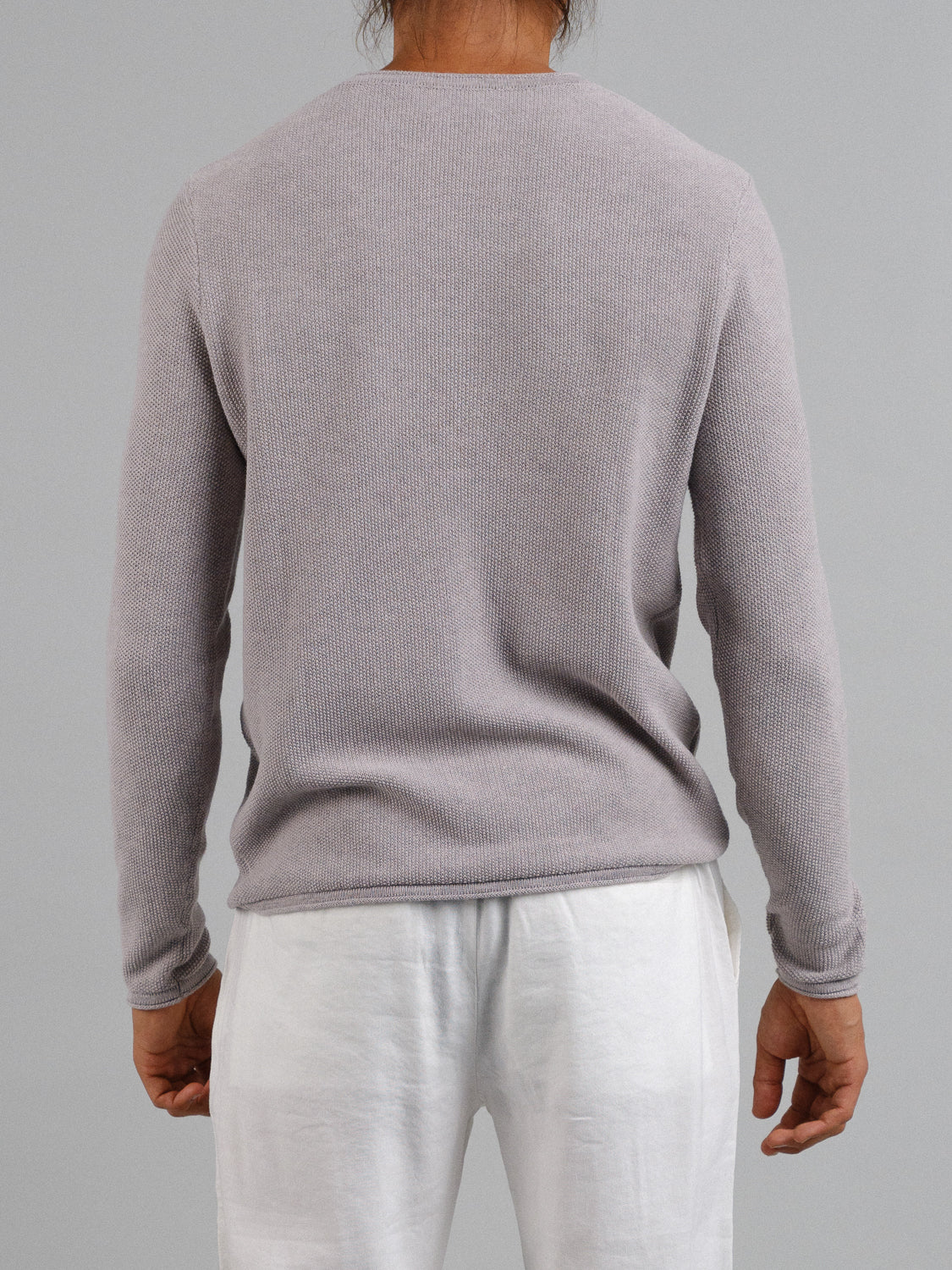 A person with long hair stands facing away, wearing an ID Menswear Madrid Rolled Neck Sweater Tee in light gray and white pants against a gray background.