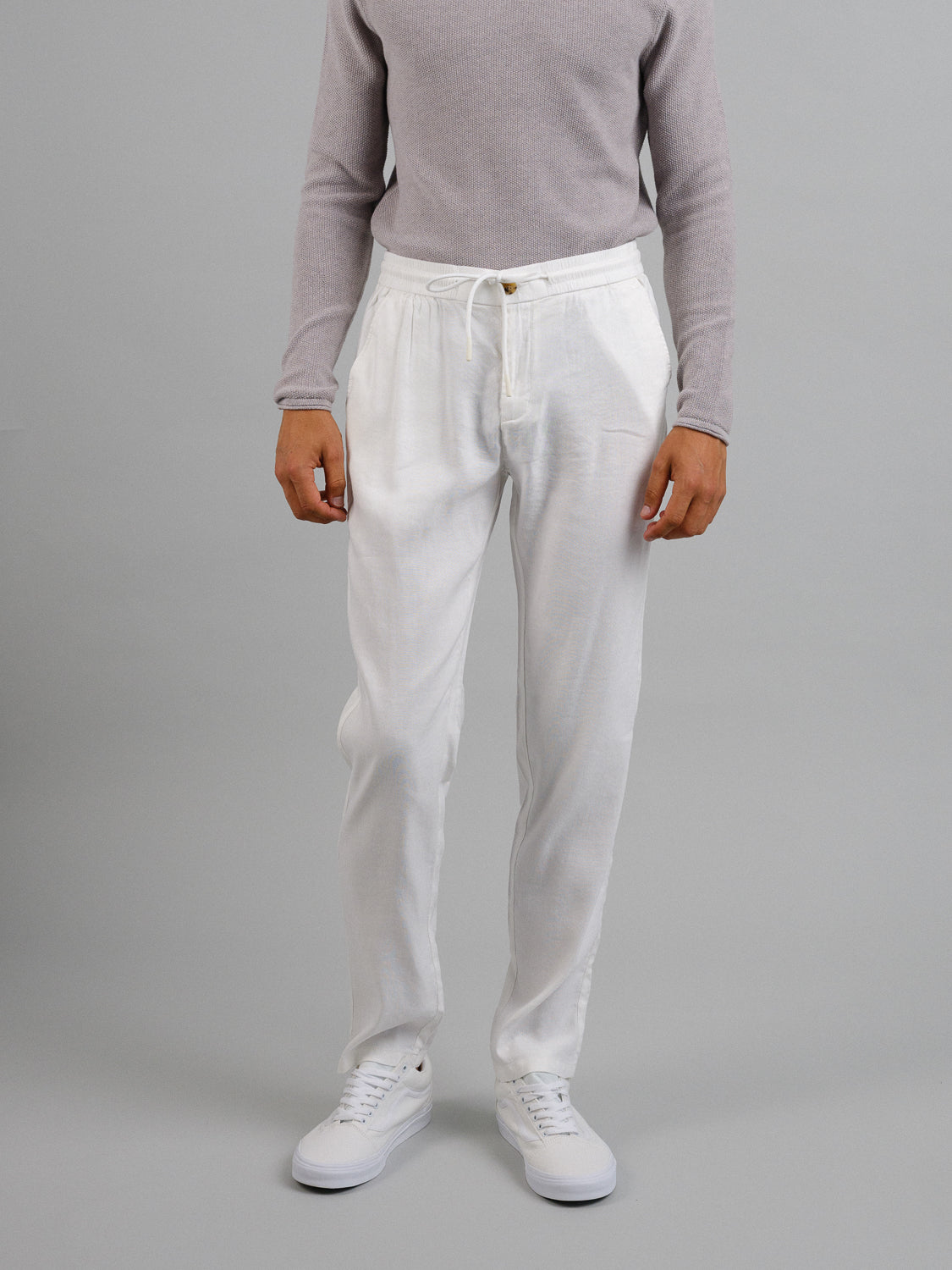 Against a plain gray backdrop, a person wears an ID Menswear light gray sweater and Bedford23 Linen Blend Drawstring Pant featuring an elastic waistband. Their white sneakers perfectly complement the outfit, adding casual elegance, but their face is hidden from view.