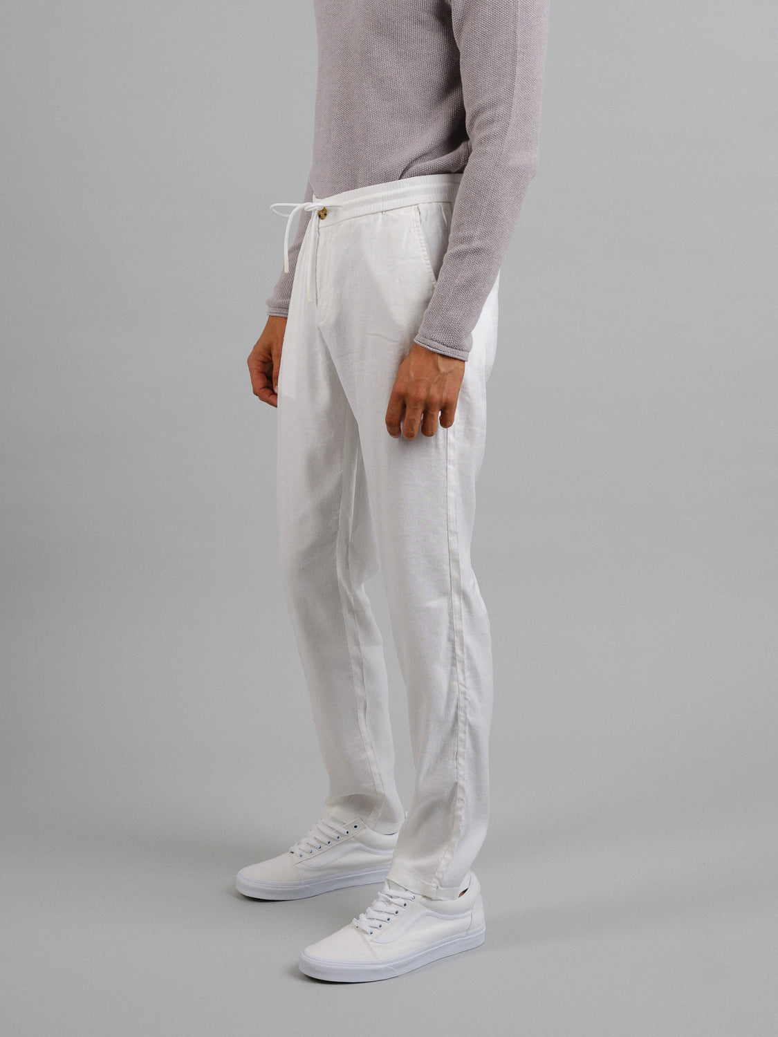 A person stands sideways wearing a gray sweater and white ID Menswear Bedford23 Linen Blend Drawstring Pants, paired with white sneakers. The neutral gray background enhances the outfits effortless style and comfort.