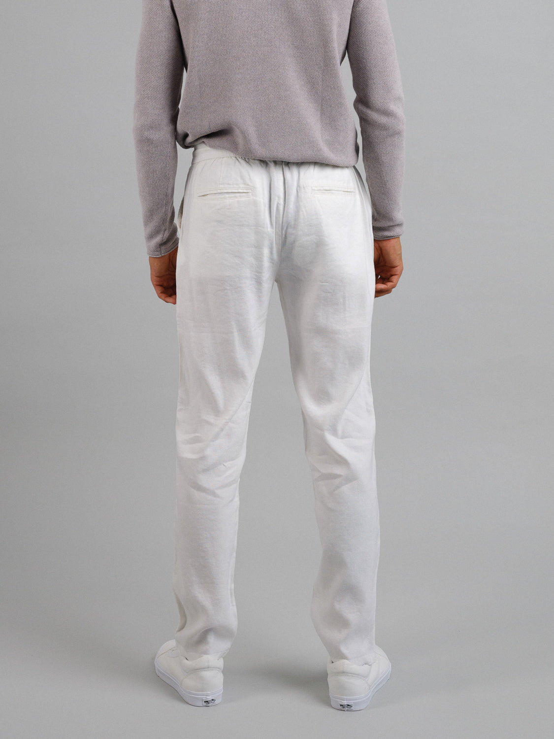 A person wearing a light gray sweater and ID Menswears Bedford23 Linen Blend Drawstring Pant stands with their back to the camera. The white pants are lightweight and comfortable, featuring an elastic drawstring waistband, complemented by white shoes, against a plain, neutral background.