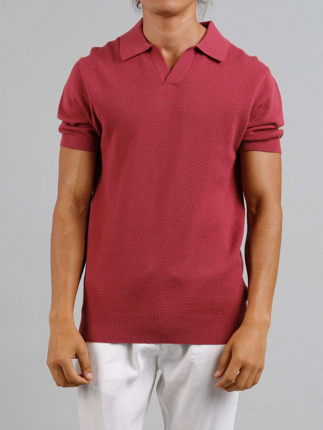 A person in an ID Menswear Malaga Knitted Cotton Polo, short-sleeved and red with a textured pattern, pairs it with white pants against a plain gray background, effortlessly exuding timeless elegance with their arms relaxed at their sides.