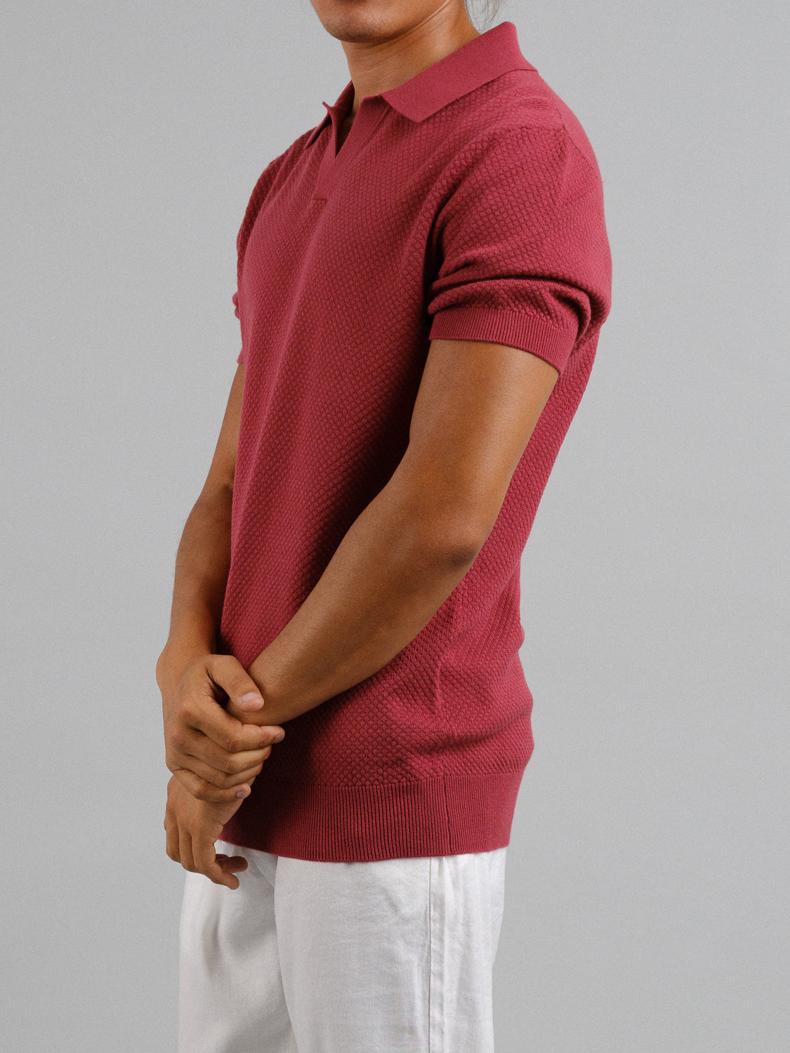 Person in an ID Menswear Malaga Knitted Cotton Polo, featuring a textured red design and short sleeves, stands with arms crossed in white pants against a plain gray background, radiating timeless elegance.