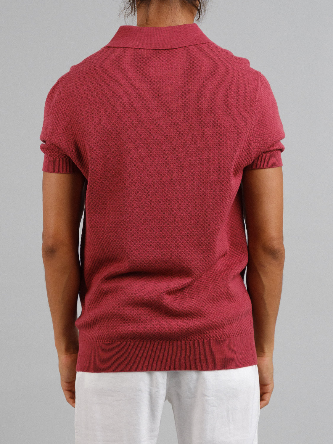 Against a simple gray wall, a person stands with their back to the camera, showcasing timeless elegance in white pants and the ID Menswear’s Malaga Knitted Cotton Polo, a textured red short-sleeve shirt made of 100% cotton.