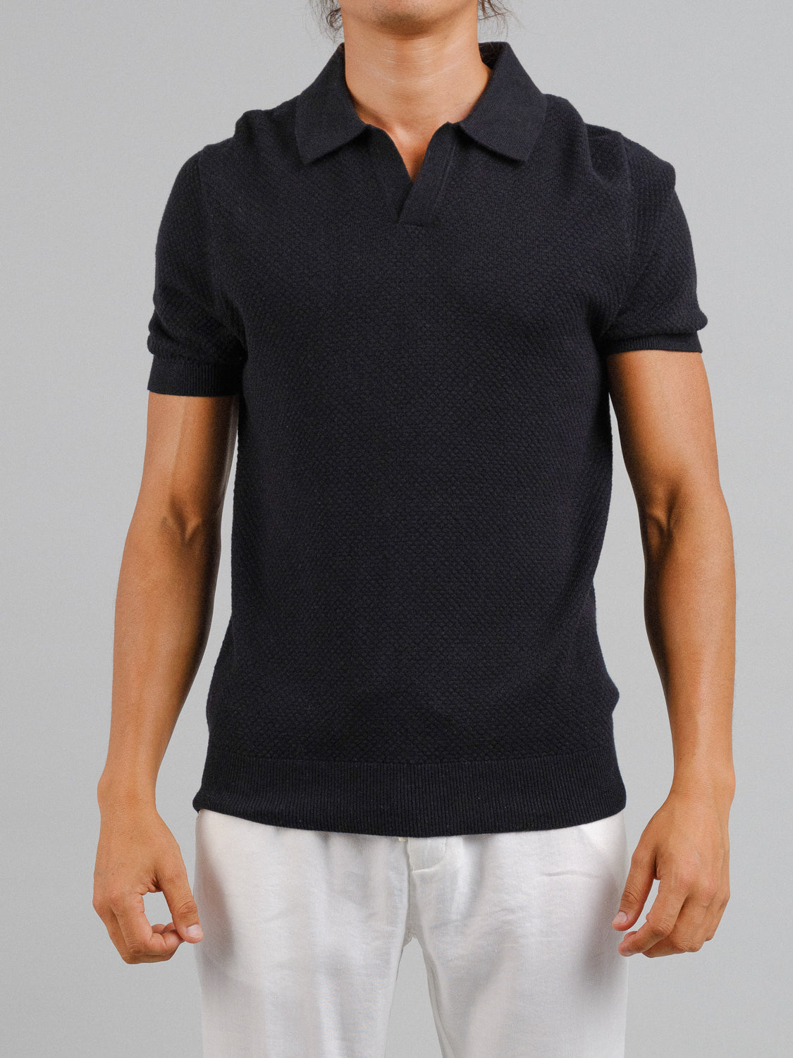 A person in a black short-sleeve Malaga Knitted Cotton Polo by ID Menswear and white pants stands against a gray background. The shirts timeless textured pattern complements their relaxed demeanor, with hands at their sides. Their head isnt visible.
