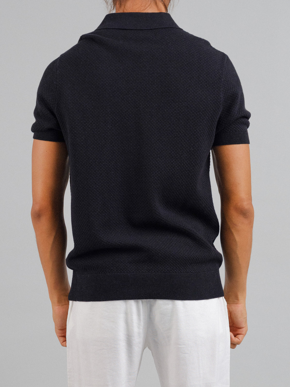 A person is seen from behind wearing a dark, short-sleeved Malaga Knitted Cotton Polo from ID Menswear and light, smooth pants. The polos plain texture adds timeless elegance against the neutral gray background.
