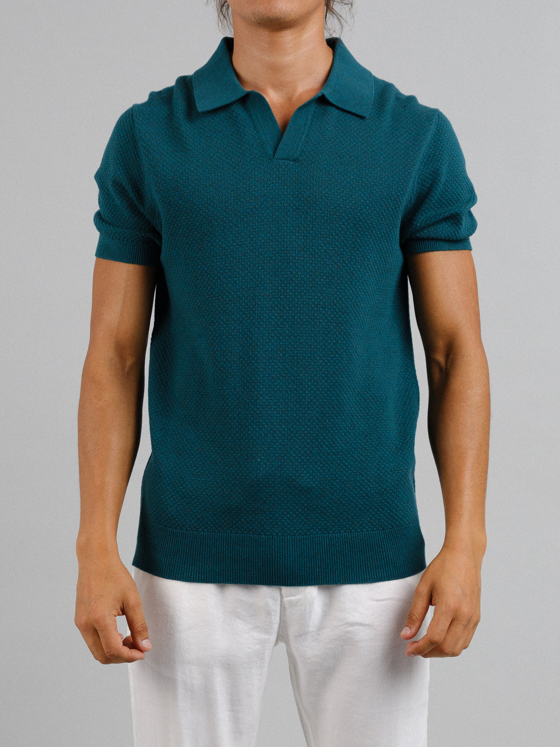 Against a gray backdrop, a person wears ID Menswears Malaga Knitted Cotton Polo in teal. This short-sleeve shirt, made from 100% cotton, boasts a textured pattern and classic collar as their arms rest by their sides.
