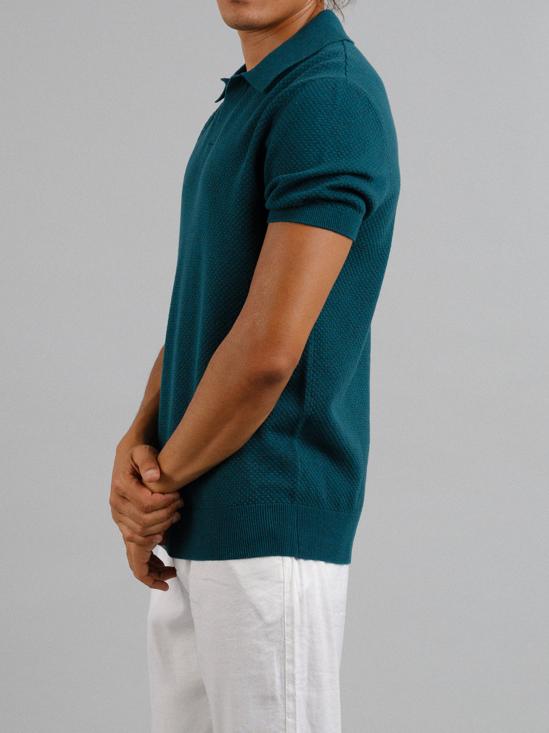 A person wearing an ID Menswear Malaga Knitted Cotton Polo in teal exudes timeless elegance against a plain backdrop, with hands clasped. Theyre paired with white pants, and the image is cropped at the head.