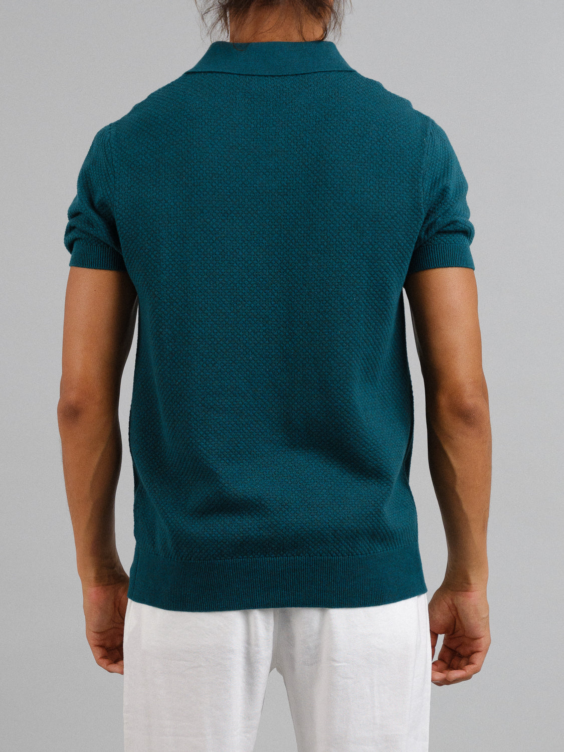 A person with long hair is seen from the back wearing an ID Menswear Malaga Knitted Cotton Polo in teal and white pants. The short-sleeved, 100% cotton shirt exudes timeless elegance against a plain gray backdrop.
