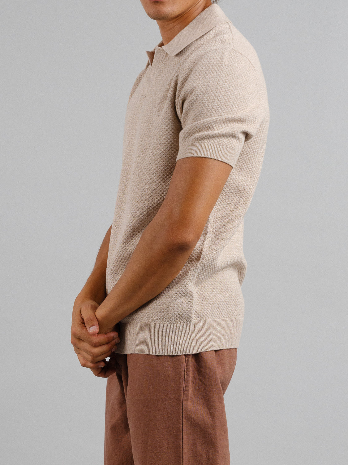 Against a gray backdrop, a person in brown pants and ID Menswears Malaga Knitted Cotton Polo, cream-colored and crafted from 100% cotton, stands sideways with hands clasped. The ensemble highlights timeless elegance through texture and color.