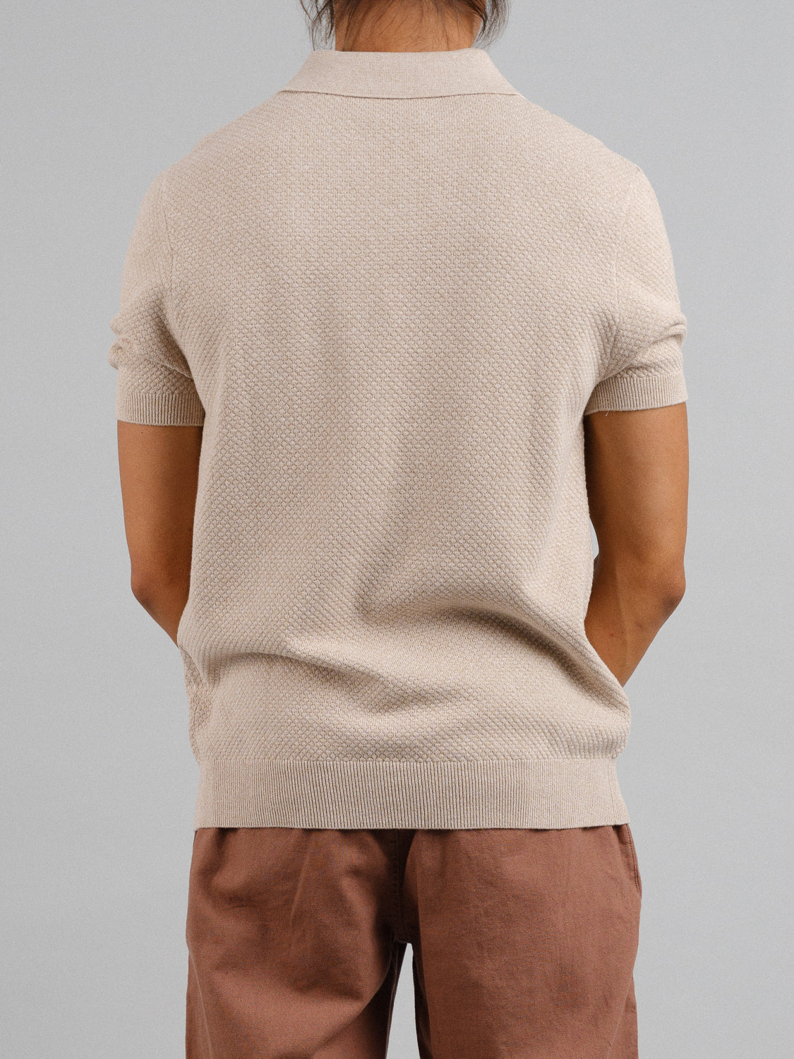 A person is seen from behind in an ID Menswear Malaga Knitted Cotton Polo, exuding timeless elegance with its textured pattern. Paired with brown pants against a neutral gray background, this beige, short-sleeved 100% cotton sweater offers a refined style.