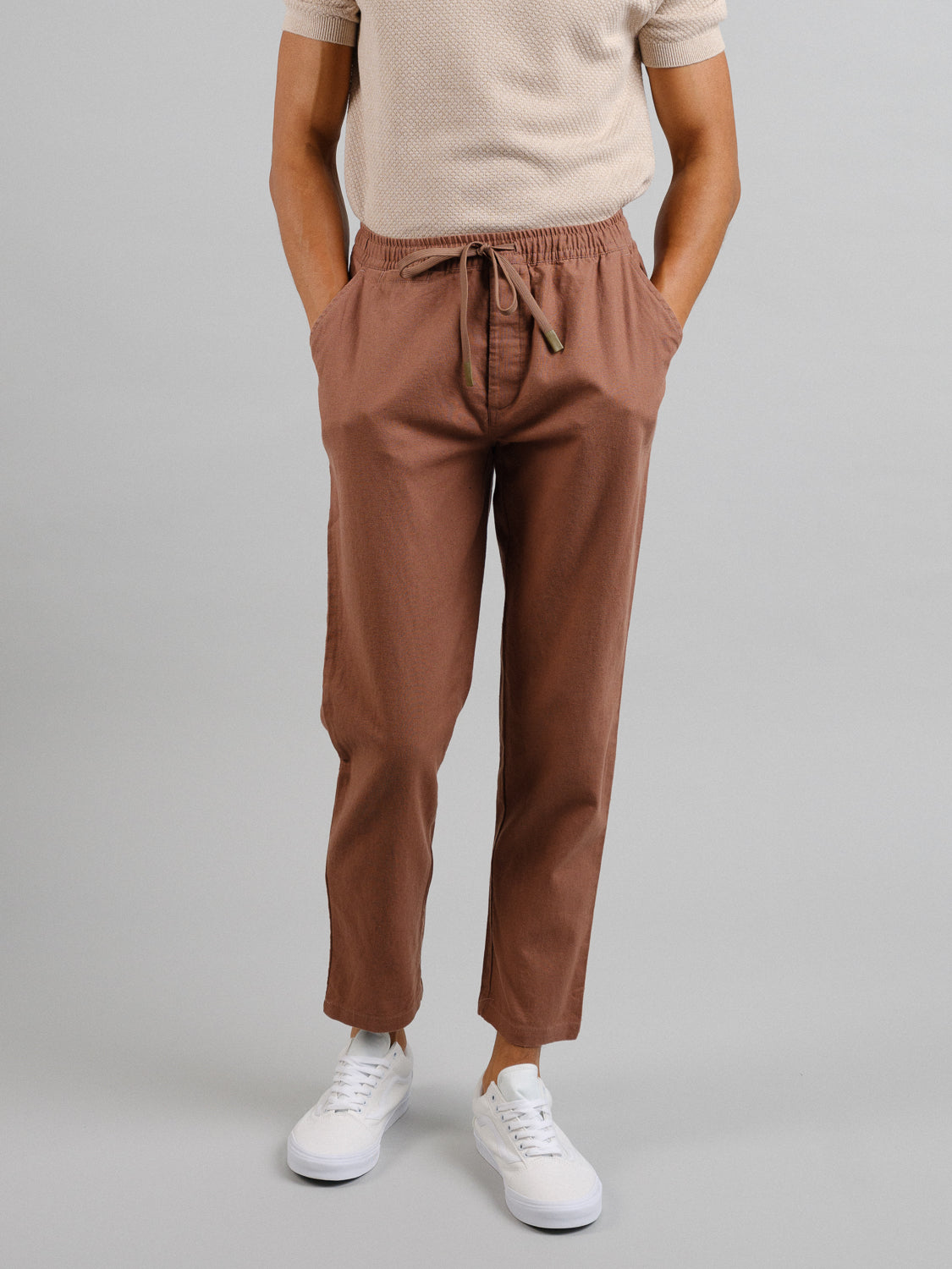 A person is wearing ID Menswears Laguna Linen Cotton Drawstring Pants, recognized for their relaxed crop fit. Theyre paired with a short-sleeved beige shirt and white sneakers against a plain gray backdrop. Only the lower half is visible, hands resting in pockets within the light brown ensemble.