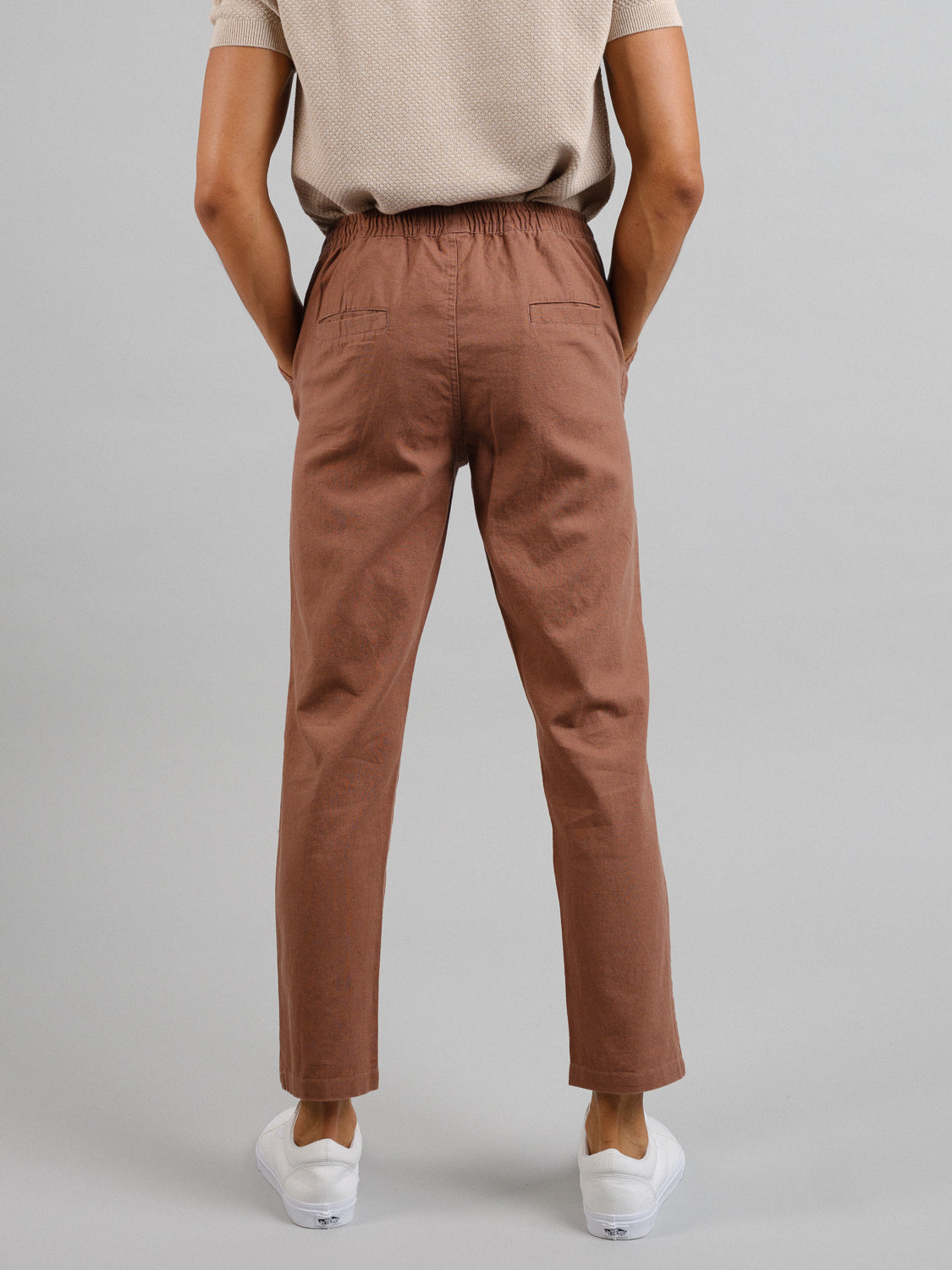 A person in a beige shirt and brown ID Menswear Laguna Linen Cotton Drawstring Pant stands with their back to the camera. The relaxed crop fit pants are paired with white sneakers against a plain gray background.