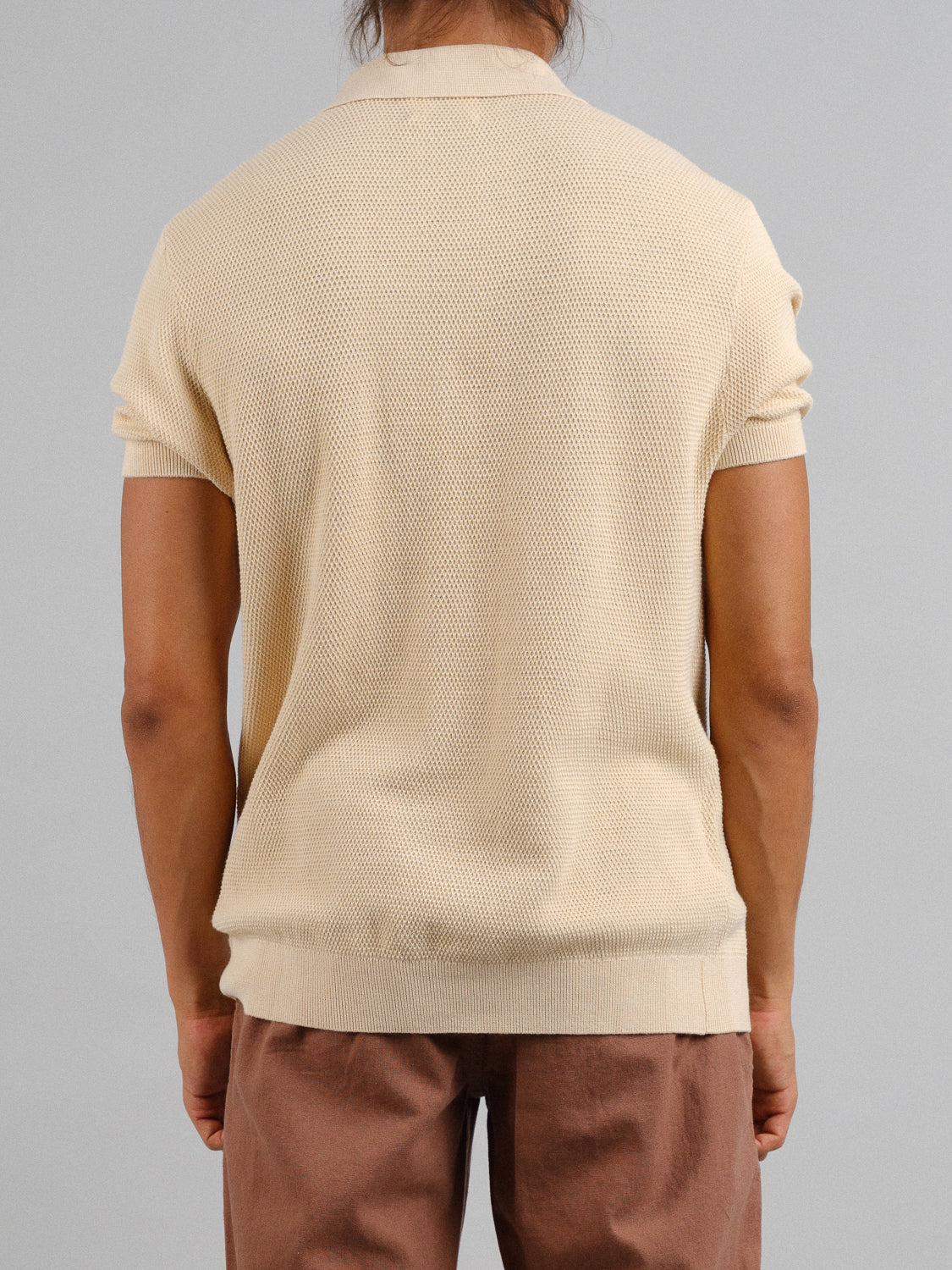 The image shows a person from behind wearing the ID Menswear Monaco Knitted Cotton Polo, which is a short-sleeved, beige top made of 100% cotton, paired with brown pants against a plain gray background.