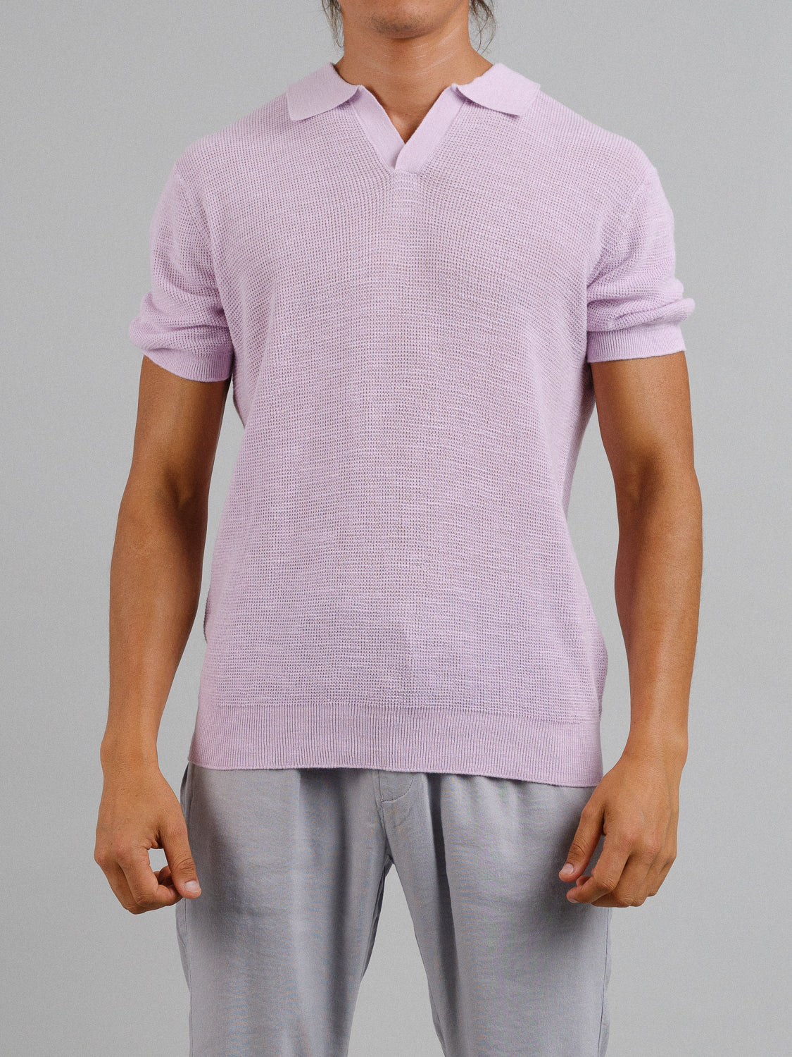 Wearing ID Menswears Ankara Linen Blend Waffle Knit Polo in a breathable light pink, paired with light gray pants, they pose against a plain gray background, showcasing the perfect warm-weather style.