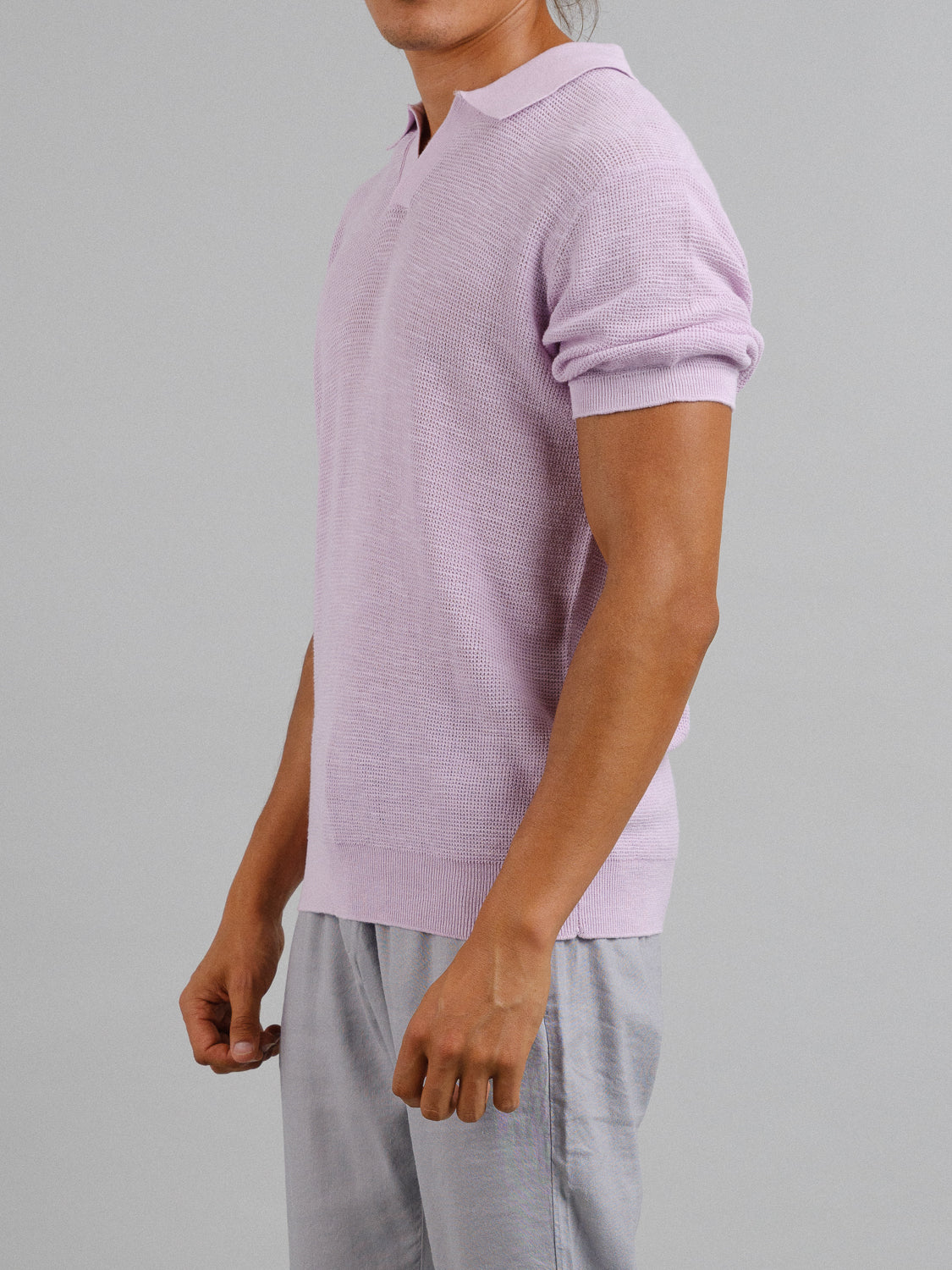 The individual sports an ID Menswear Ankara Linen Blend Waffle Knit Polo, a light purple, lightweight and breathable short-sleeve sweater with a collar, paired with light gray pants. Relaxed arms by their sides against a plain gray background create an ideal warm weather look.