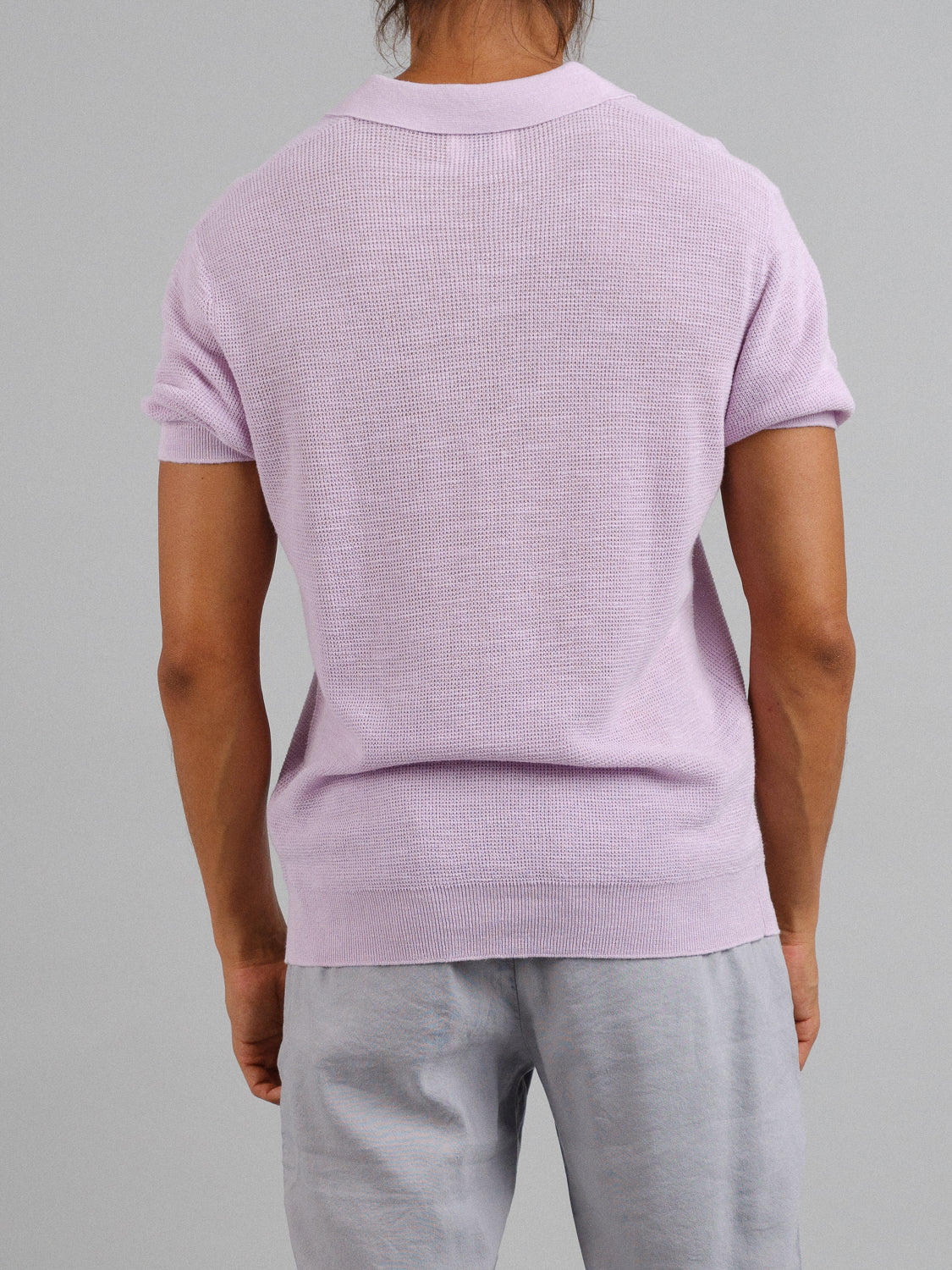 A person with long hair stands against a gray background, wearing the ID Menswear Ankara Linen Blend Waffle Knit Polo in light purple and light gray pants, ideal for warm weather. The polos lightweight, breathable fabric ensures comfort and style.