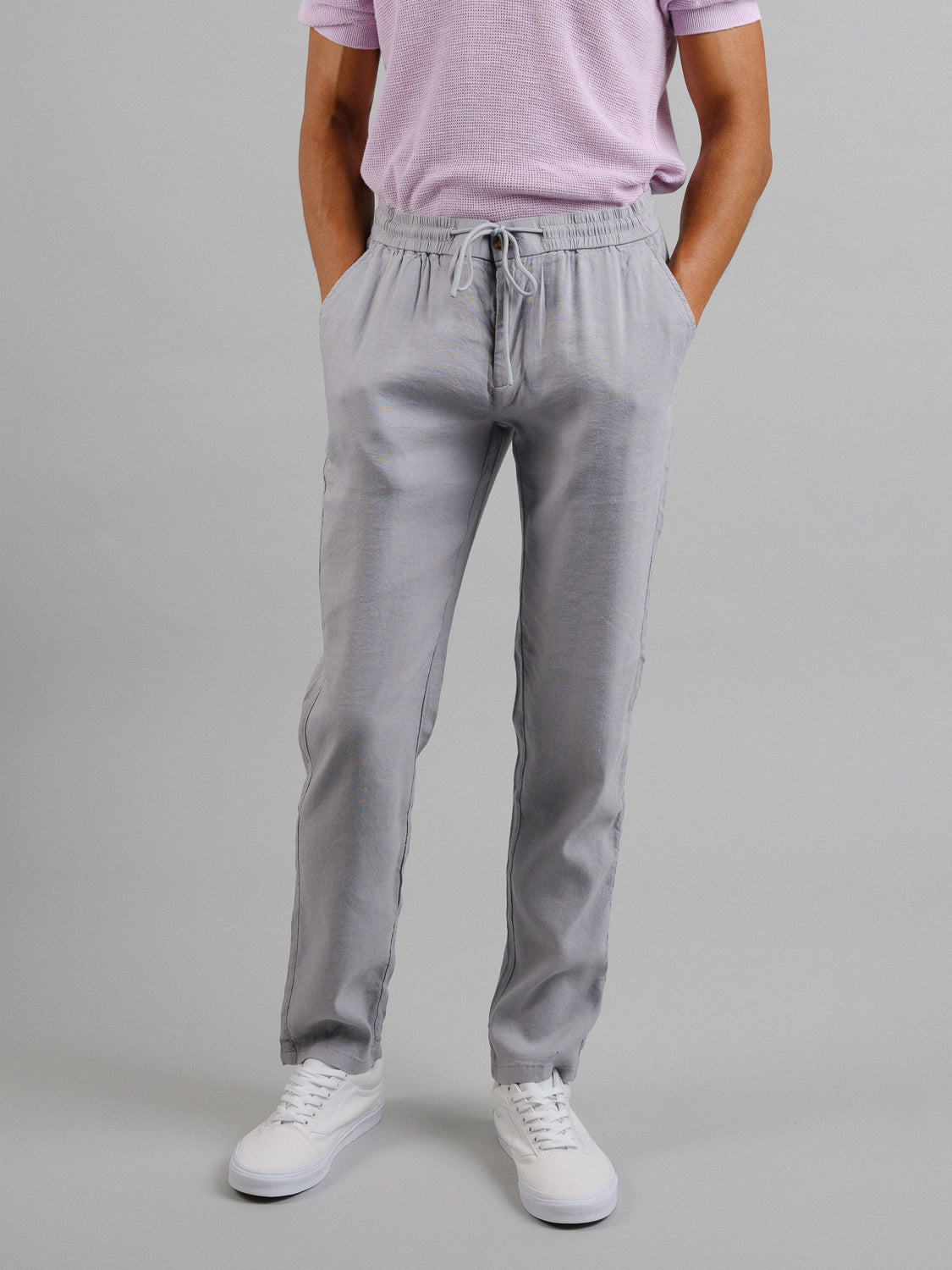 A person in a light purple t-shirt, ID Menswear Bedford23 Linen Blend Drawstring Pant with an elastic waistband, and white sneakers stands against a plain gray background. Their hands are in their pockets, and the photo is cropped at the shoulders.