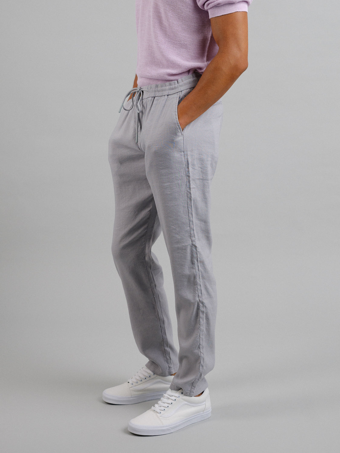 A person in light gray ID Menswear Bedford23 Linen Blend Drawstring Pants, a pink shirt, and white sneakers stands against a plain background. The comfortable pants feature an elastic drawstring waistband, with their hands casually tucked into the pockets. Their face is not visible.