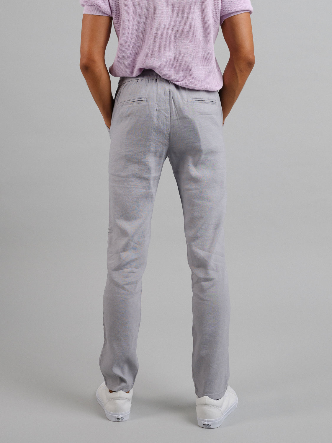 A person wears ID Menswears Bedford23 Linen Blend Drawstring Pant, featuring an elastic waistband, and a light purple shirt. They stand against a gray background, paired with classic white sneakers for an effortless fit.