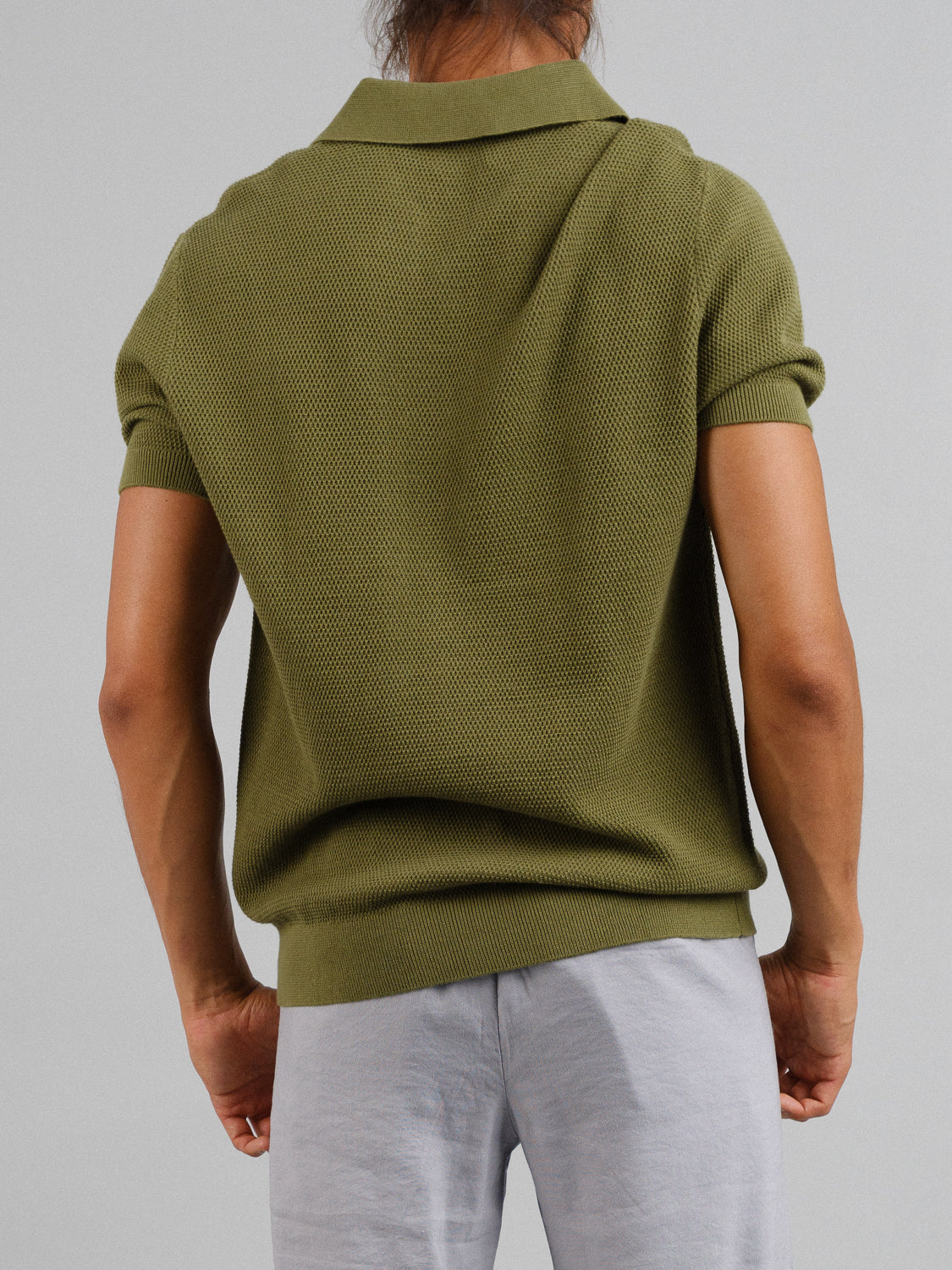 Rear view of a person wearing an ID Menswear Monaco Knitted Cotton Polo in green with short sleeves and light gray pants, both made from 100% cotton, set against a plain gray background.
