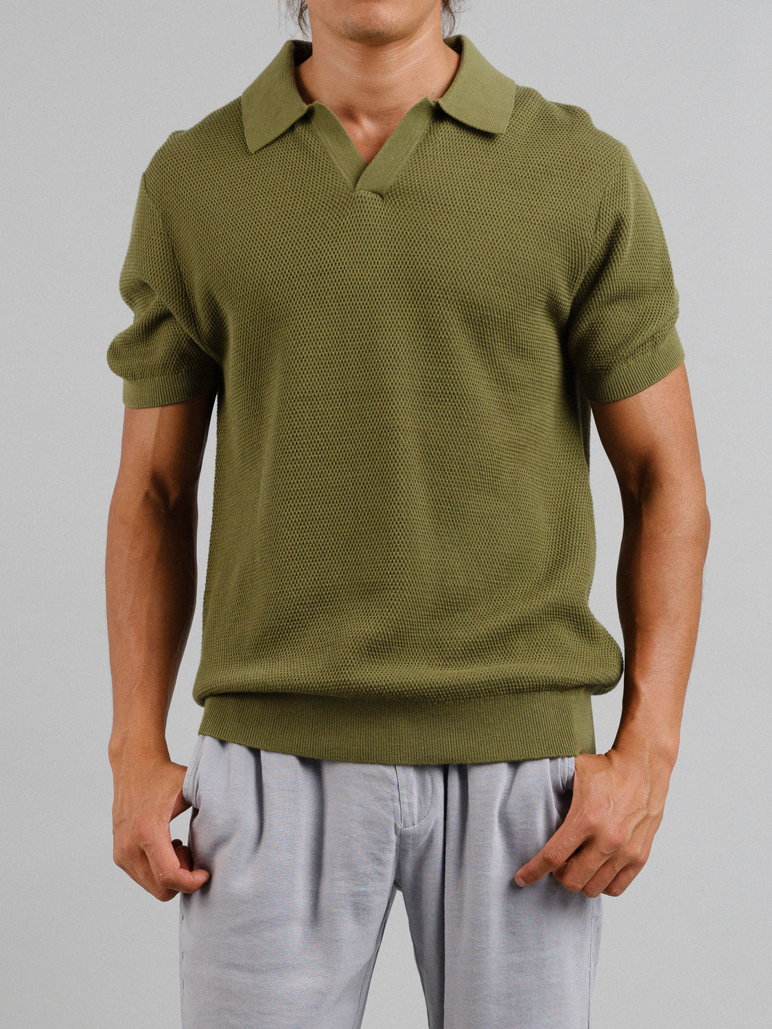 A person in an ID Menswear Monaco Knitted Cotton Polo, short-sleeved and olive green with a spread collar, paired with light gray pants. The image highlights the torso, arms relaxed at the sides, set against a plain gray background.