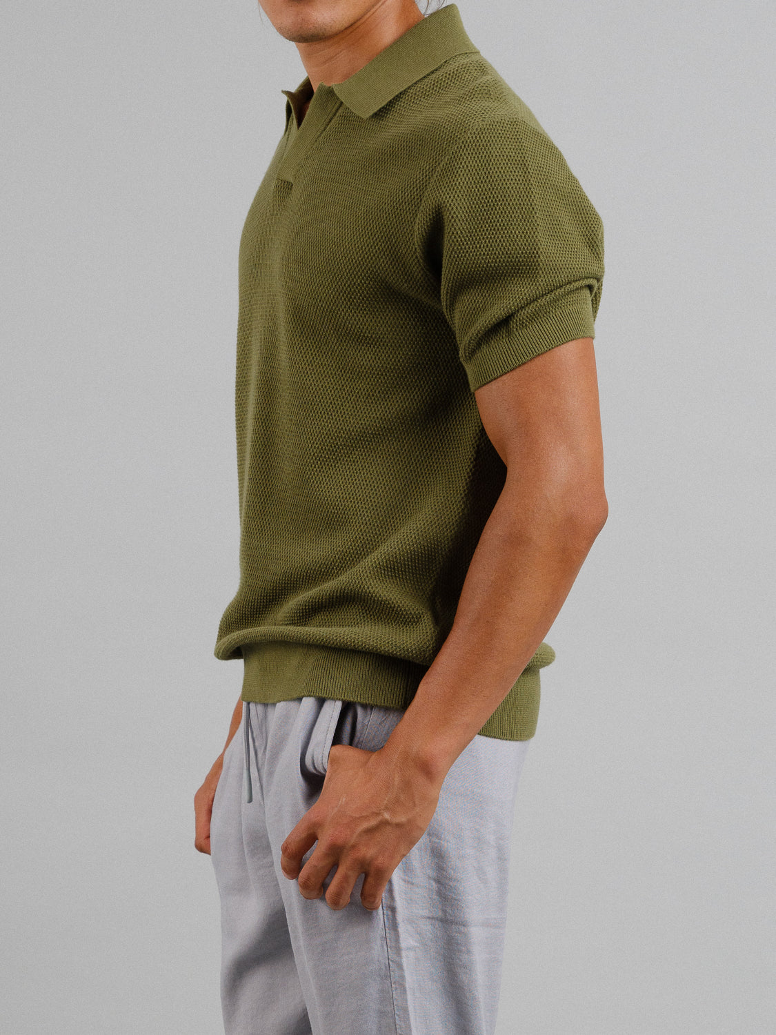 A person in an olive green Monaco Knitted Cotton Polo by ID Menswear and light gray pants stands sideways against a plain gray background, with their face hidden.