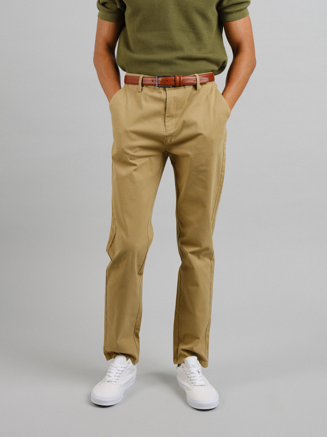 A person in a green shirt, ID Menswears Bushwick Twill Stretch Chino in khaki, a brown belt, and white sneakers stands with hands in pockets against a plain background, the image showing the outfit from shoulders to feet.