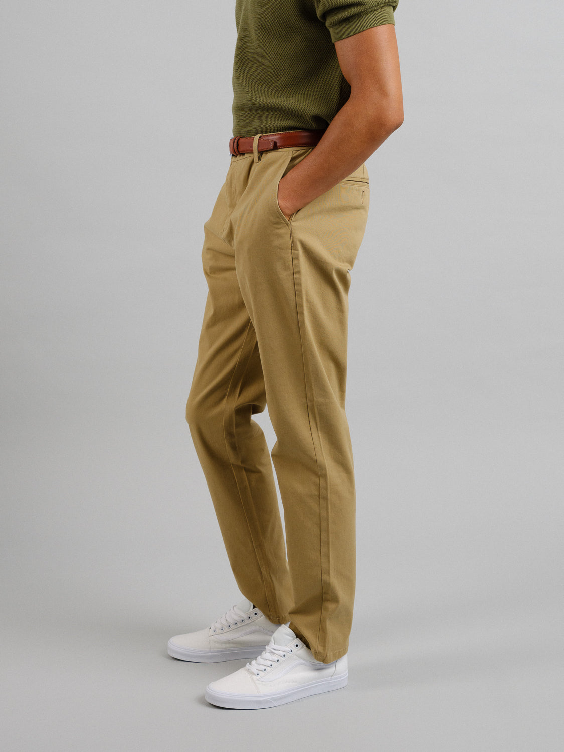 Against a plain backdrop, a person wears a green shirt with beige ID Menswear Bushwick Twill Stretch Chino trousers, secured by a brown belt. White sneakers complete the look as their left hand rests in the pocket. The persons face is not shown.