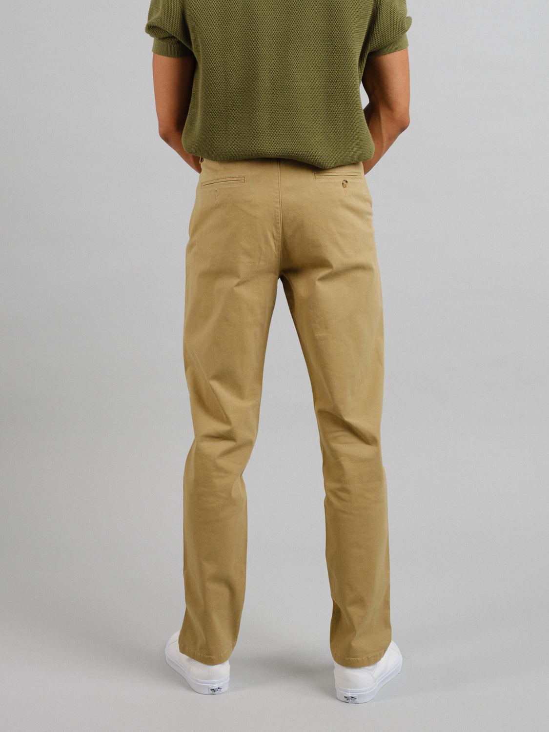 A person stands with their back to the camera, wearing a short-sleeved olive green shirt, white sneakers, and ID Menswears Bushwick Twill Stretch Chino. The background is plain and light gray.