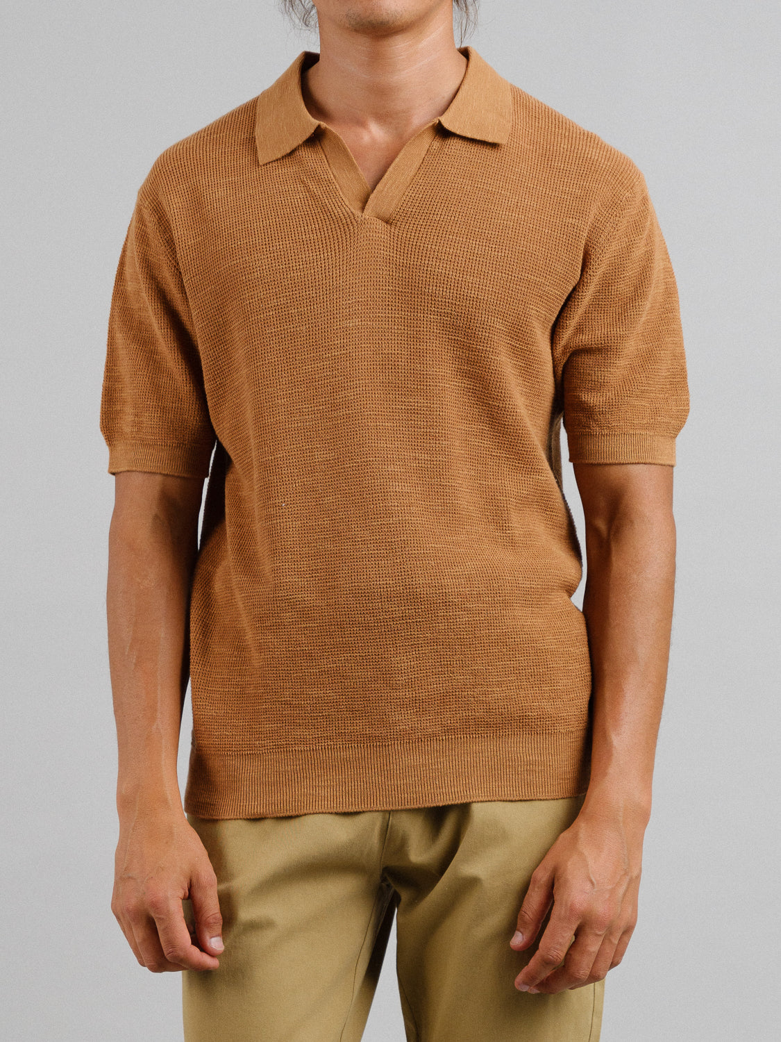 A person stands against a gray backdrop wearing ID Menswears Ankara Linen Blend Waffle Knit Polo in brown with tan pants. The short-sleeve polo, with its lightweight and breathable design, offers a textured pattern and relaxed fit. Their face is not visible.
