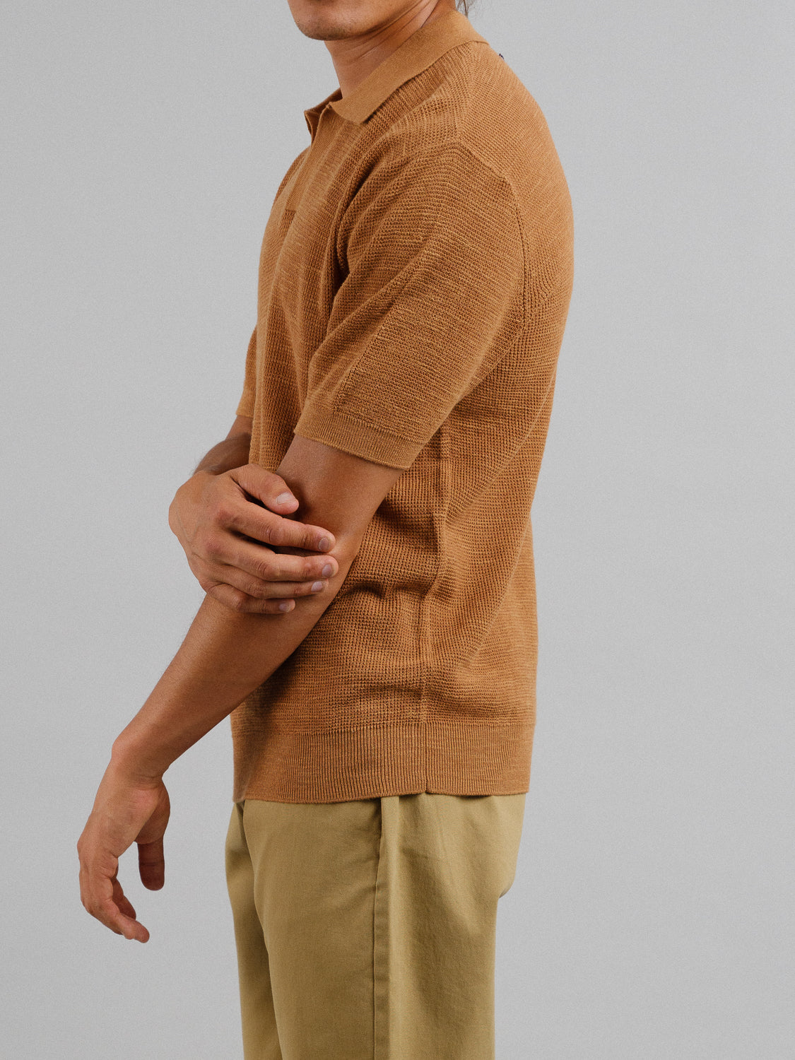 A person stands sideways against a gray backdrop wearing an Ankara Linen Blend Waffle Knit Polo by ID Menswear in tan, with short sleeves and brown pants, arms crossed loosely, shown from the waist up. Ideal for warm weather, this lightweight and breathable fabric ensures day-long comfort.