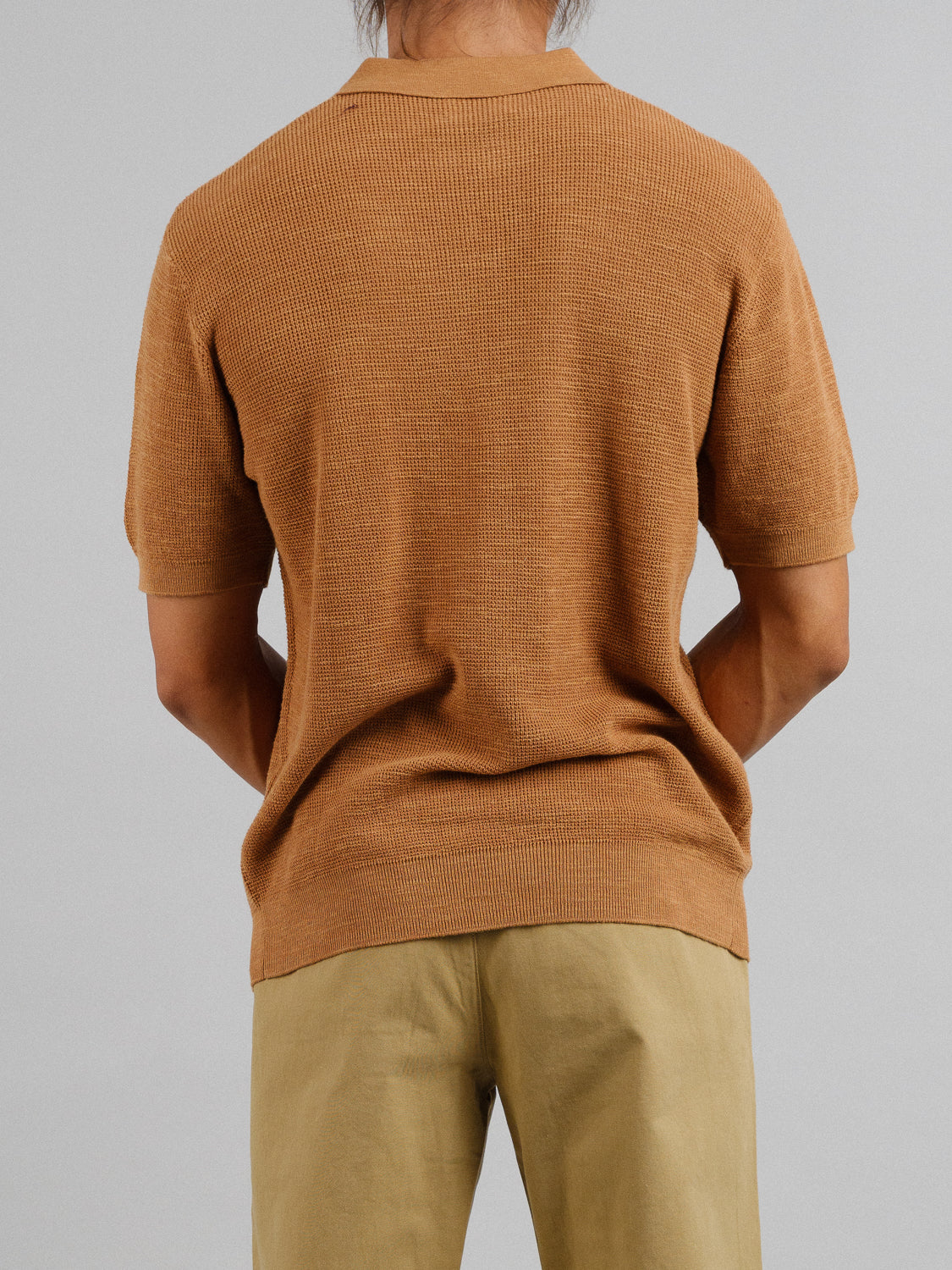 A person with long hair stands facing away from the camera, wearing an ID Menswear Ankara Linen Blend Waffle Knit Polo and beige pants. Their arms rest behind against a gray backdrop, suited for warm weather.