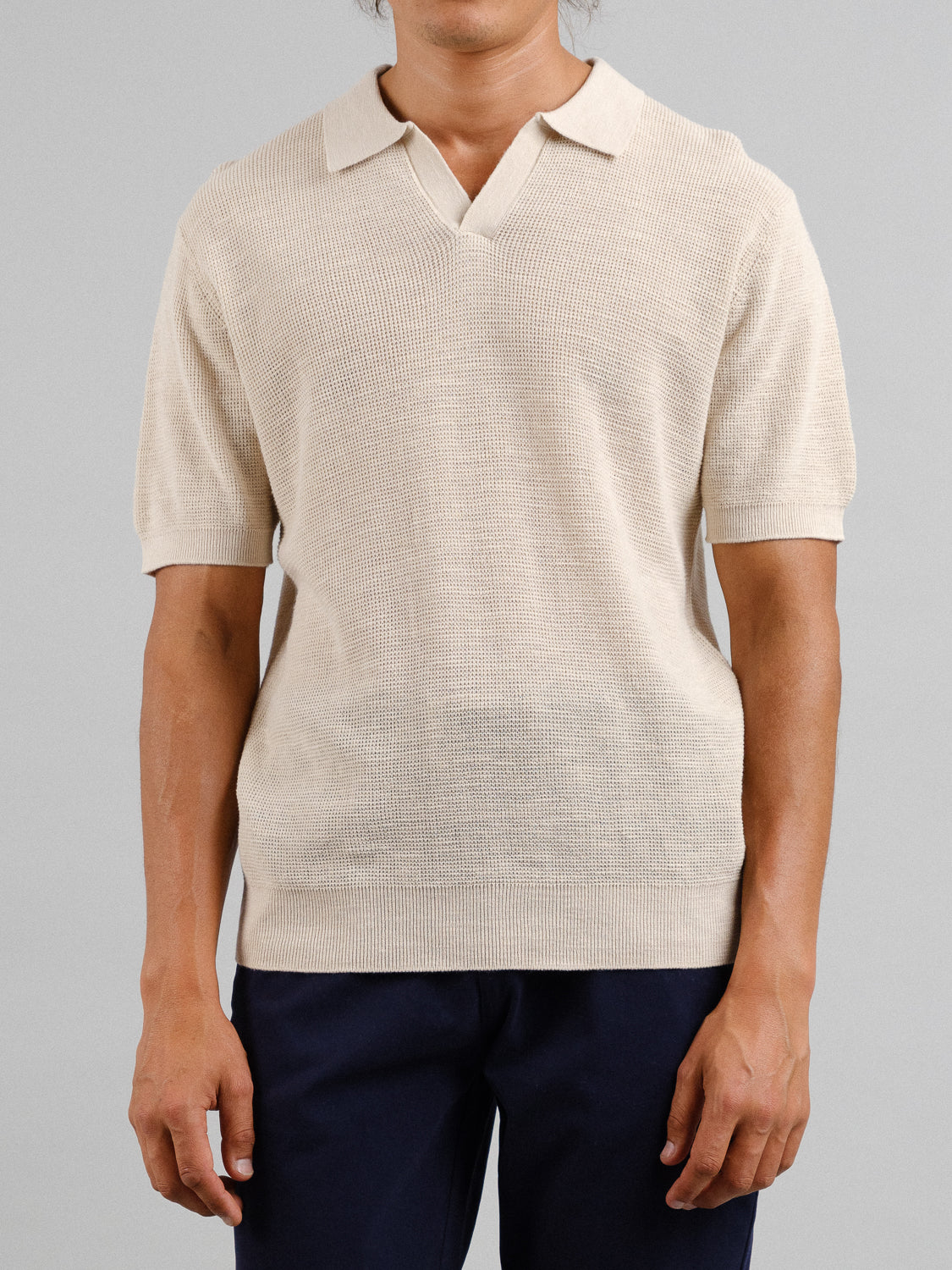 A person wears an ID Menswear Ankara Linen Blend Waffle Knit Polo, a light beige short-sleeve with a textured pattern, paired with dark navy trousers. This lightweight and breathable ensemble is perfect for warm weather. The plain gray background obscures the persons face.