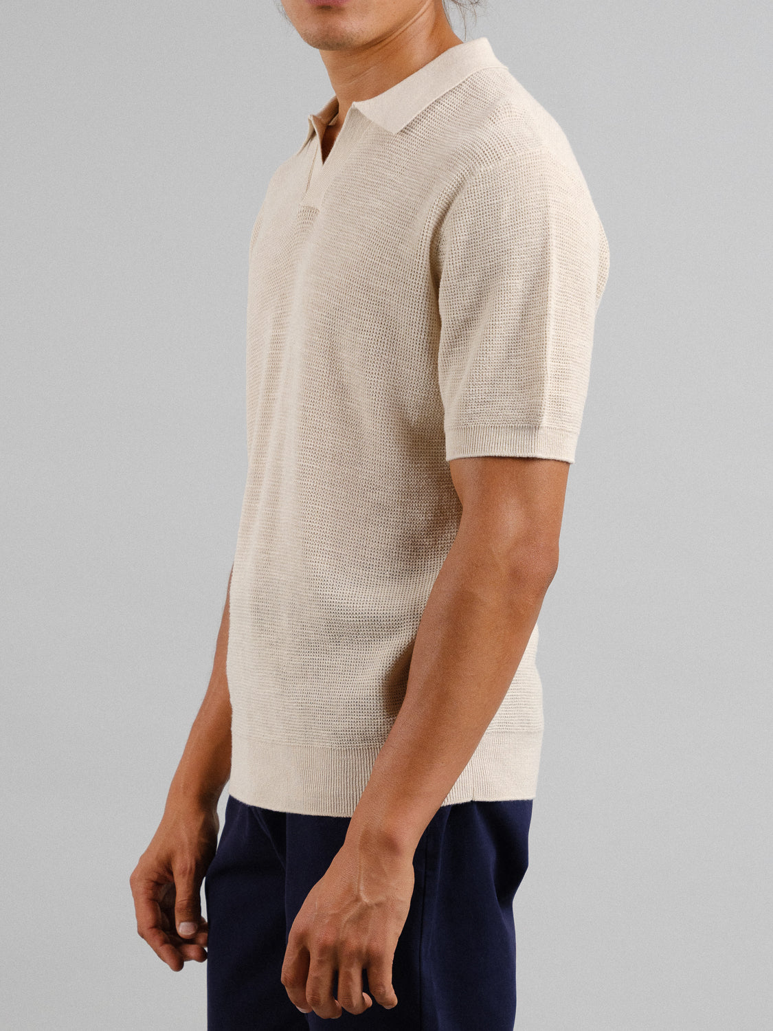 A person dons an ID Menswear Ankara Linen Blend Waffle Knit Polo in light beige paired with dark pants against a gray backdrop. The lightweight, breathable polo is ideal for warm weather, drawing attention to the stylish attire despite the head being out of view.