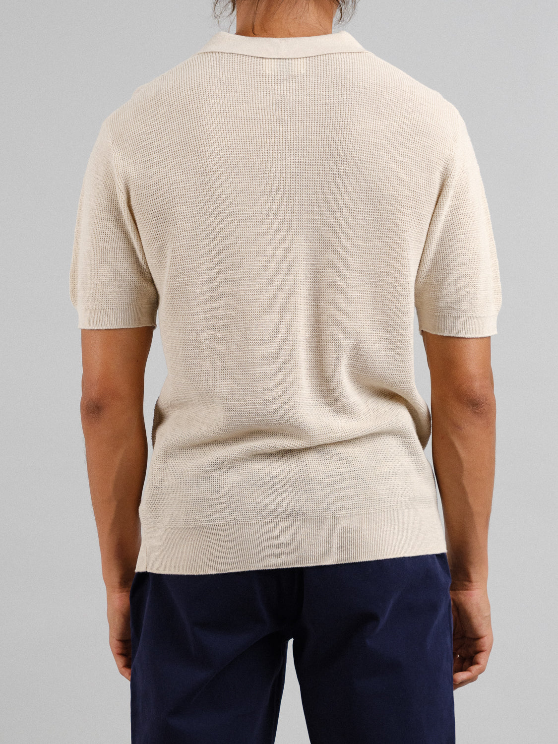 Someone stands with their back to the camera, wearing the Ankara Linen Blend Waffle Knit Polo in beige from ID Menswear and navy pants, ideal for warm weather. The lightweight, breathable fabric contrasts well with the plain light gray background.
