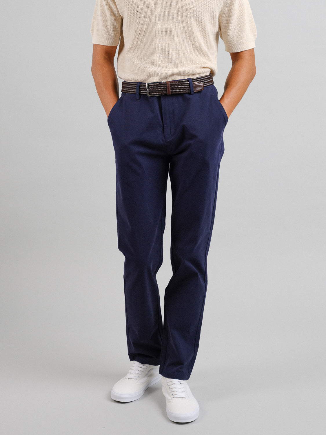 A person is wearing a beige short-sleeve shirt, ID Menswears Bushwick Twill Stretch Chino in navy, a brown belt, and white sneakers. Their hands rest in the relaxed-fit trouser pockets against a plain gray backdrop, with the upper torso not visible.