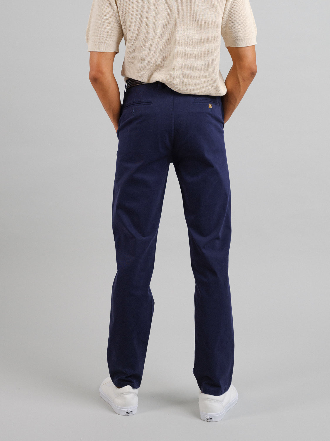 An individual is seen from behind, wearing a beige short-sleeve shirt, ID Menswears relaxed-fit Bushwick Twill Stretch Chino in navy blue, and white sneakers against a plain light gray background.