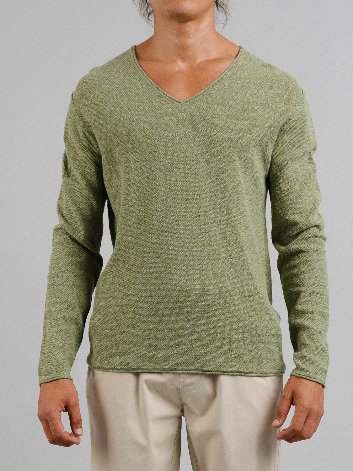 A person is wearing a light green Amalfi Deep V Knitted Top from ID Menswear paired with beige pants. The plain gray background accentuates the linen-cotton blend top’s deep V-neck, highlighting the stylish ensemble.