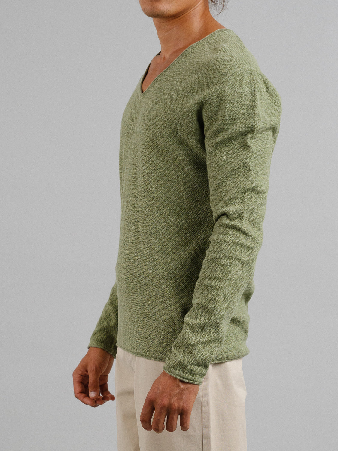 A person in a light green Amalfi Deep V Knitted Top from ID Menswear paired with beige pants is facing sideways. The neutral background accentuates the texture of the linen cotton blend sweater, though their head isnt visible in the image.